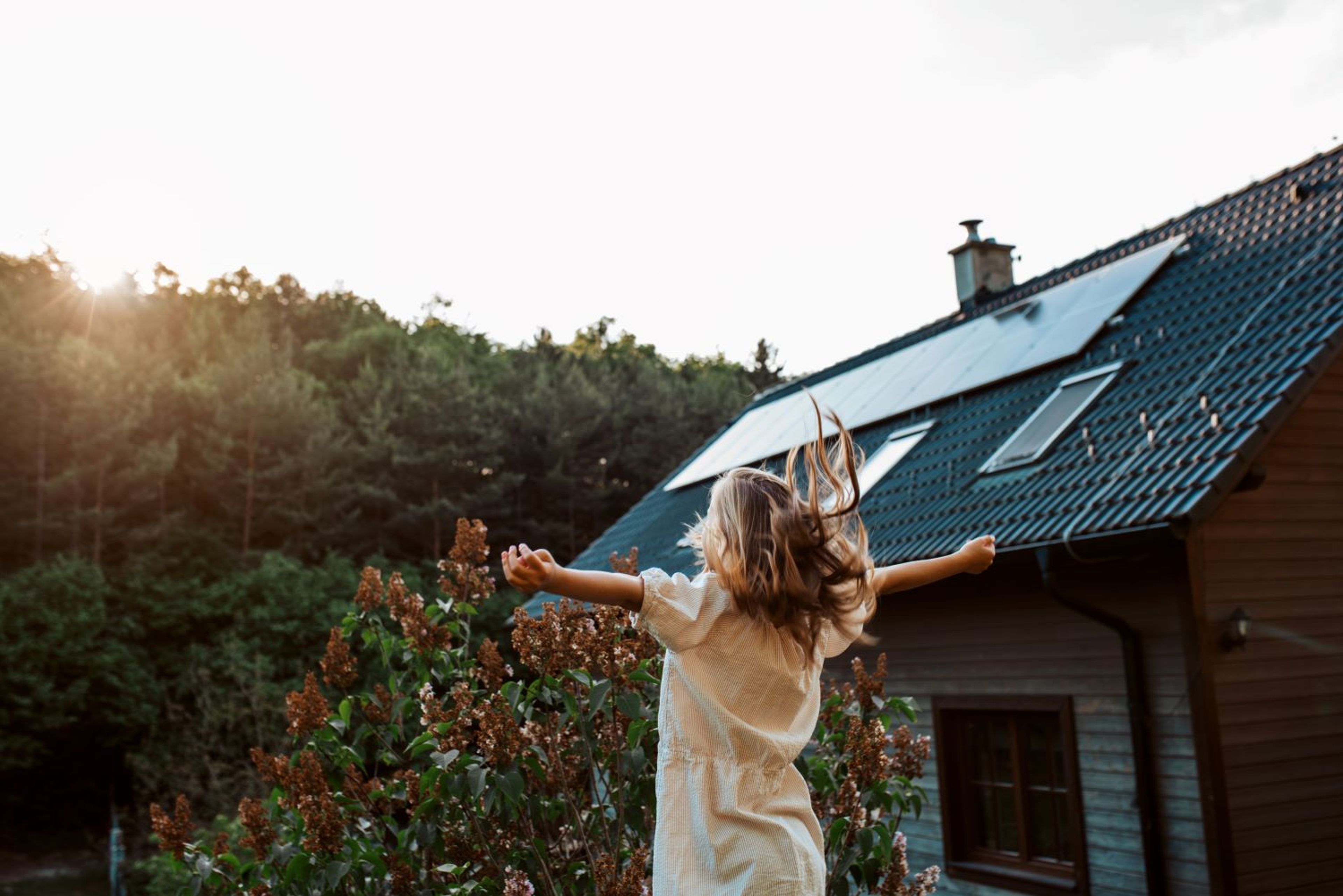 Meisje springt omhoog met lichtgeel kleedje en armen horizontaal, haar lange blonde haar wipt op. Struik en bomen in de achtergond bij een huis met zonnepanelen.
