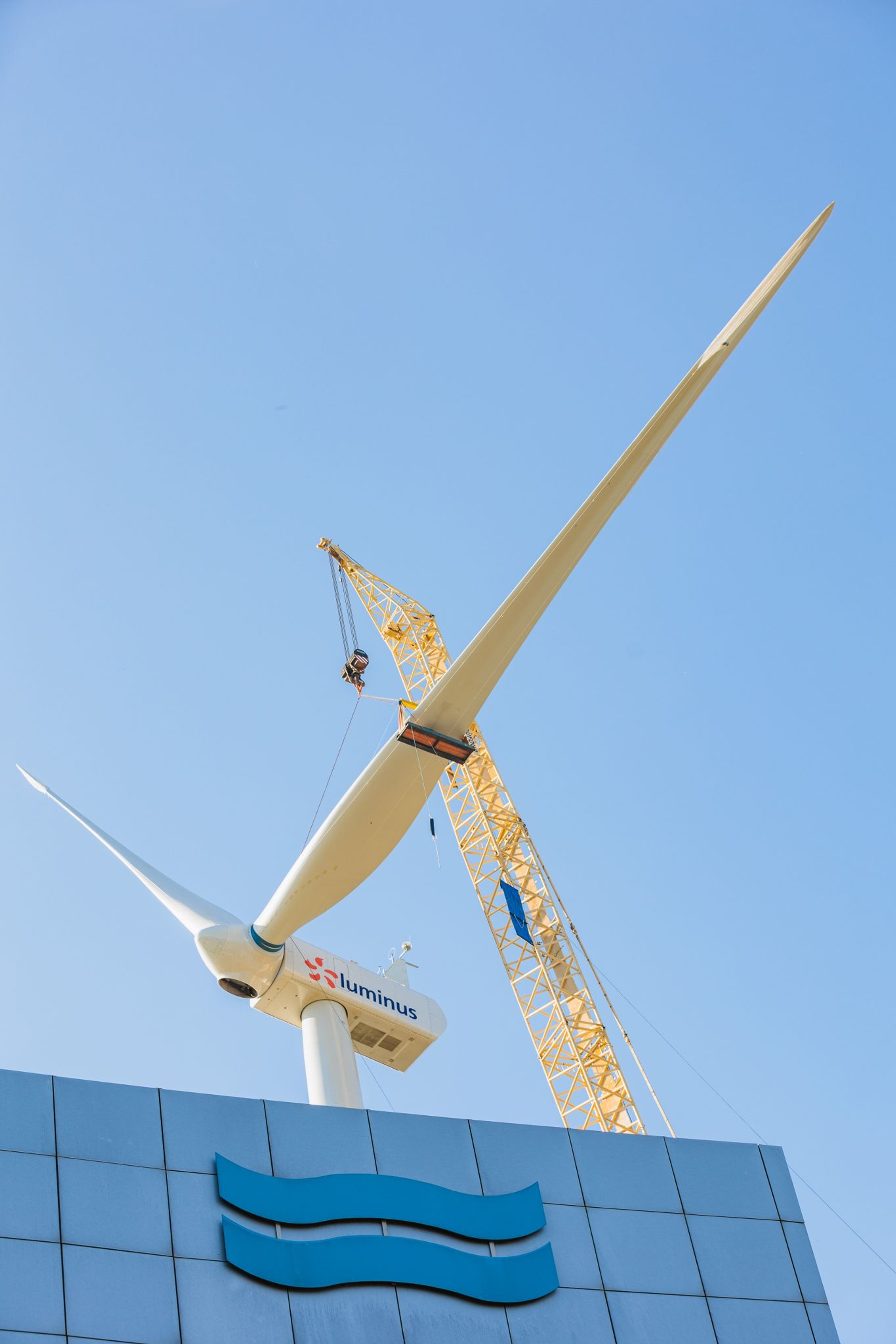 Une grande éolienne Luminus aux pales blanches se dresse à côté d’une grue jaune sous un ciel bleu clair.