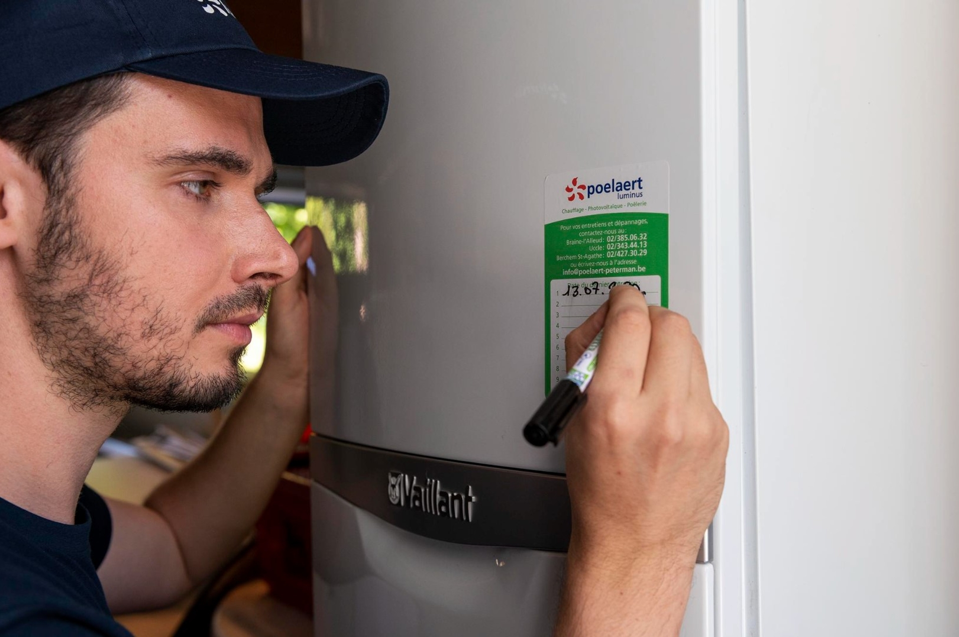 Technicien portant une casquette inscrivant une date sur une étiquette de maintenance collée sur une chaudière grise