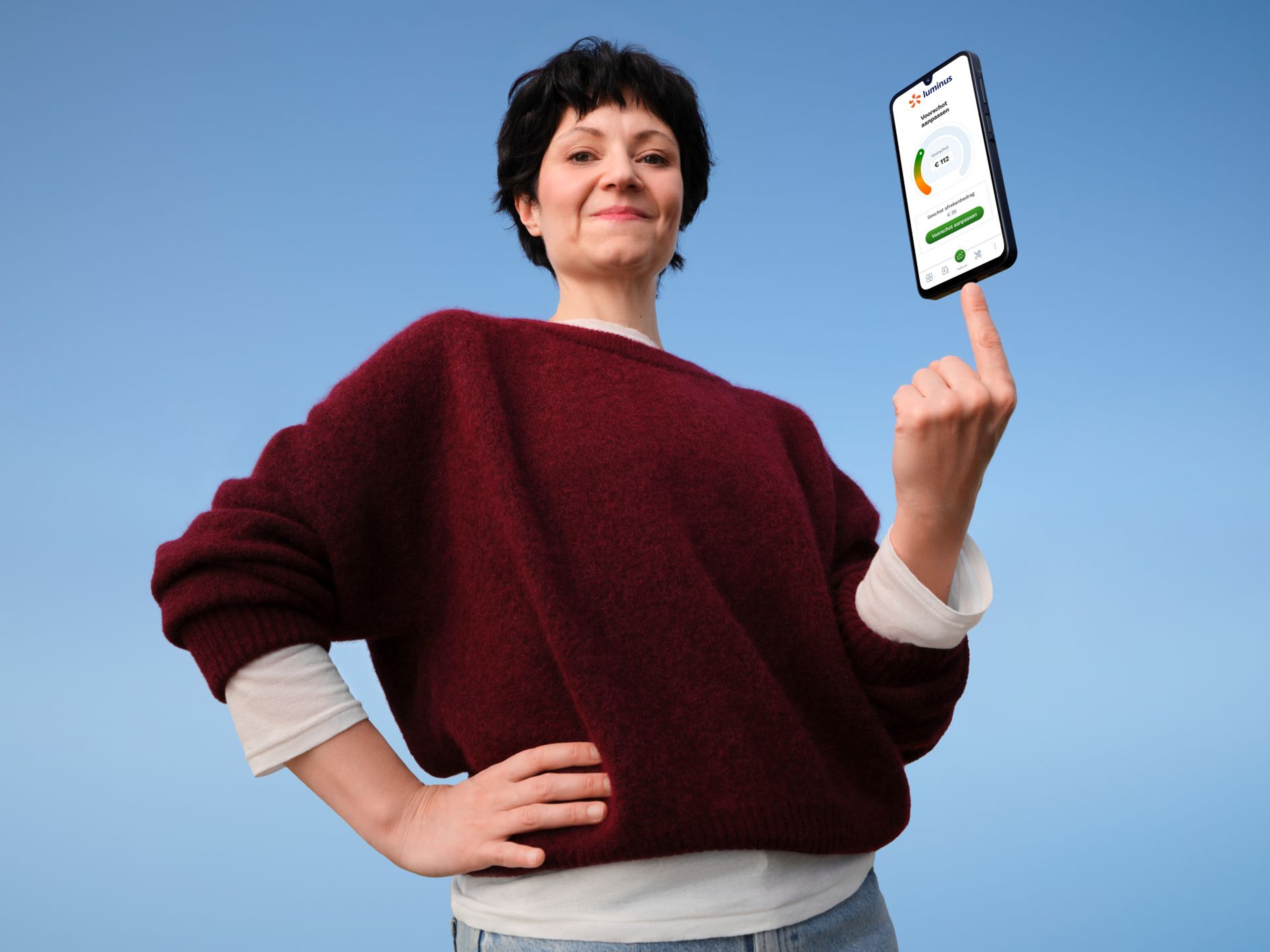 Zelfverzekerde dame met bordaux trui houdt een smartphone op 1 vinger, My Luminus dashboard op het scherm.