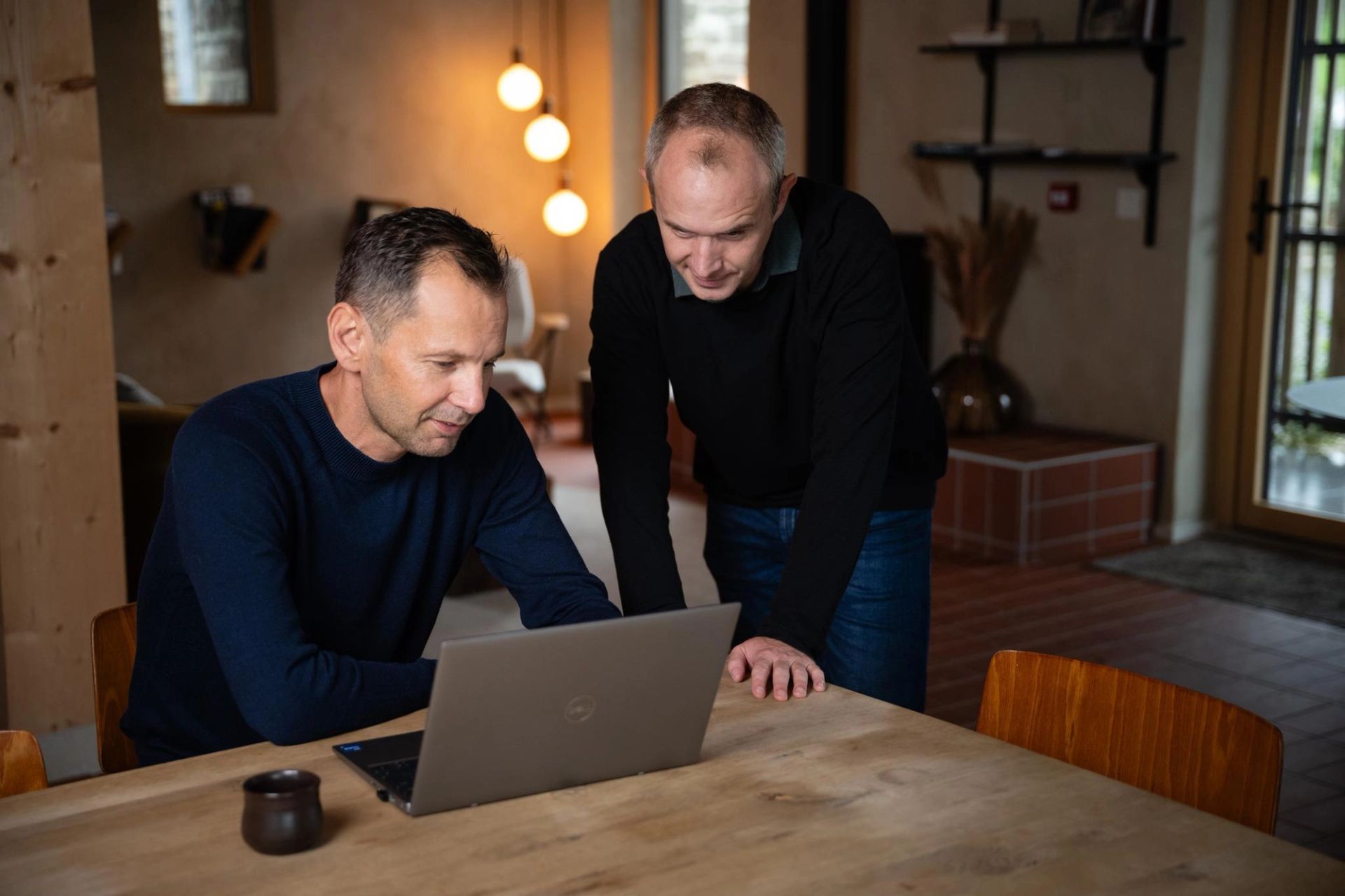 Two men in living room, one sitting, the other standing, are watching a laptop.
