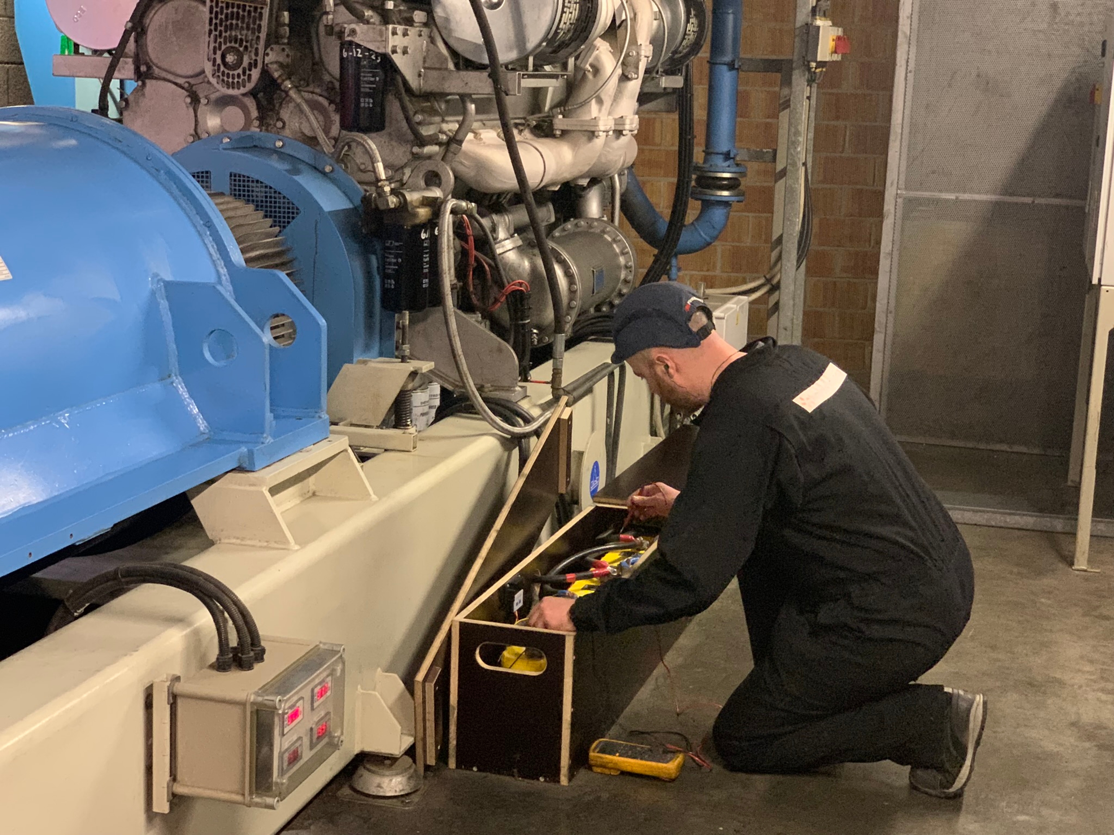 Technicien effectuant la maintenance d'un système UPS industriel, agenouillé devant un générateur dans une salle technique