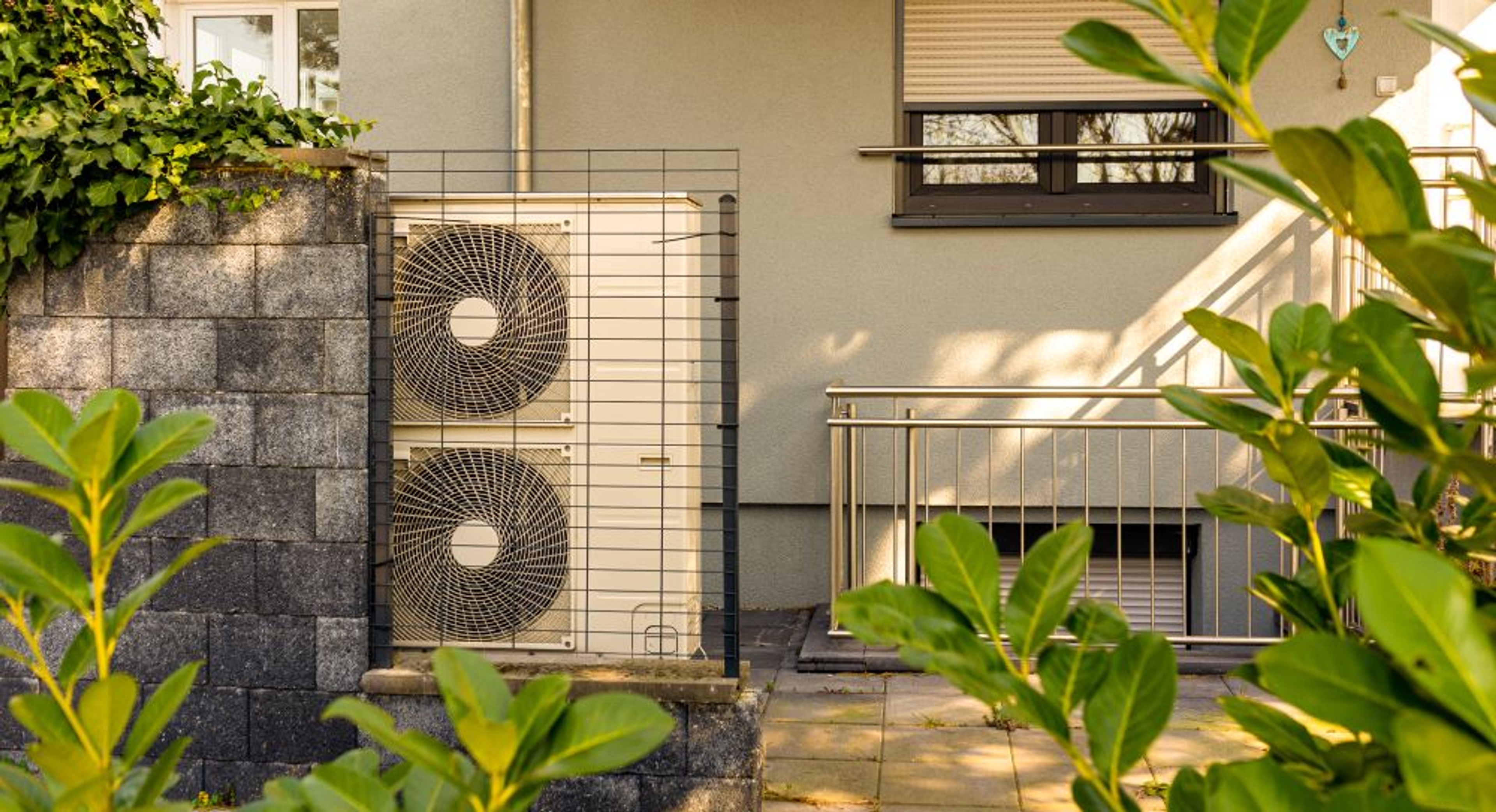 Pompe à chaleur extérieure avec deux ventilateurs installée contre le mur d’une maison, entourée de végétation et protégée par une grille métallique