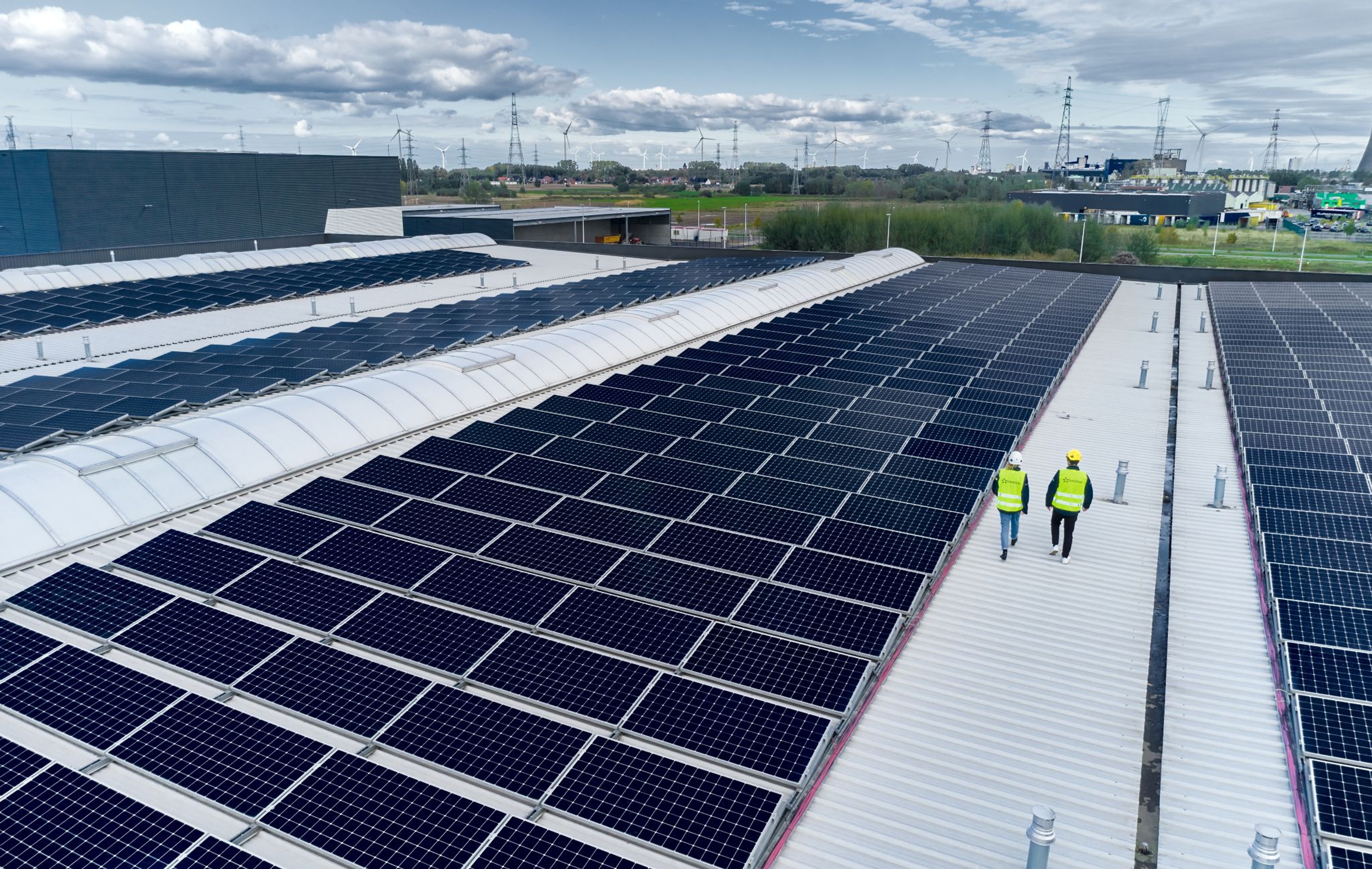 Deux techniciens en gilets de sécurité marchent sur le toit d'un bâtiment industriel couvert de panneaux solaires, avec des lignes électriques et éoliennes visibles à l'horizon