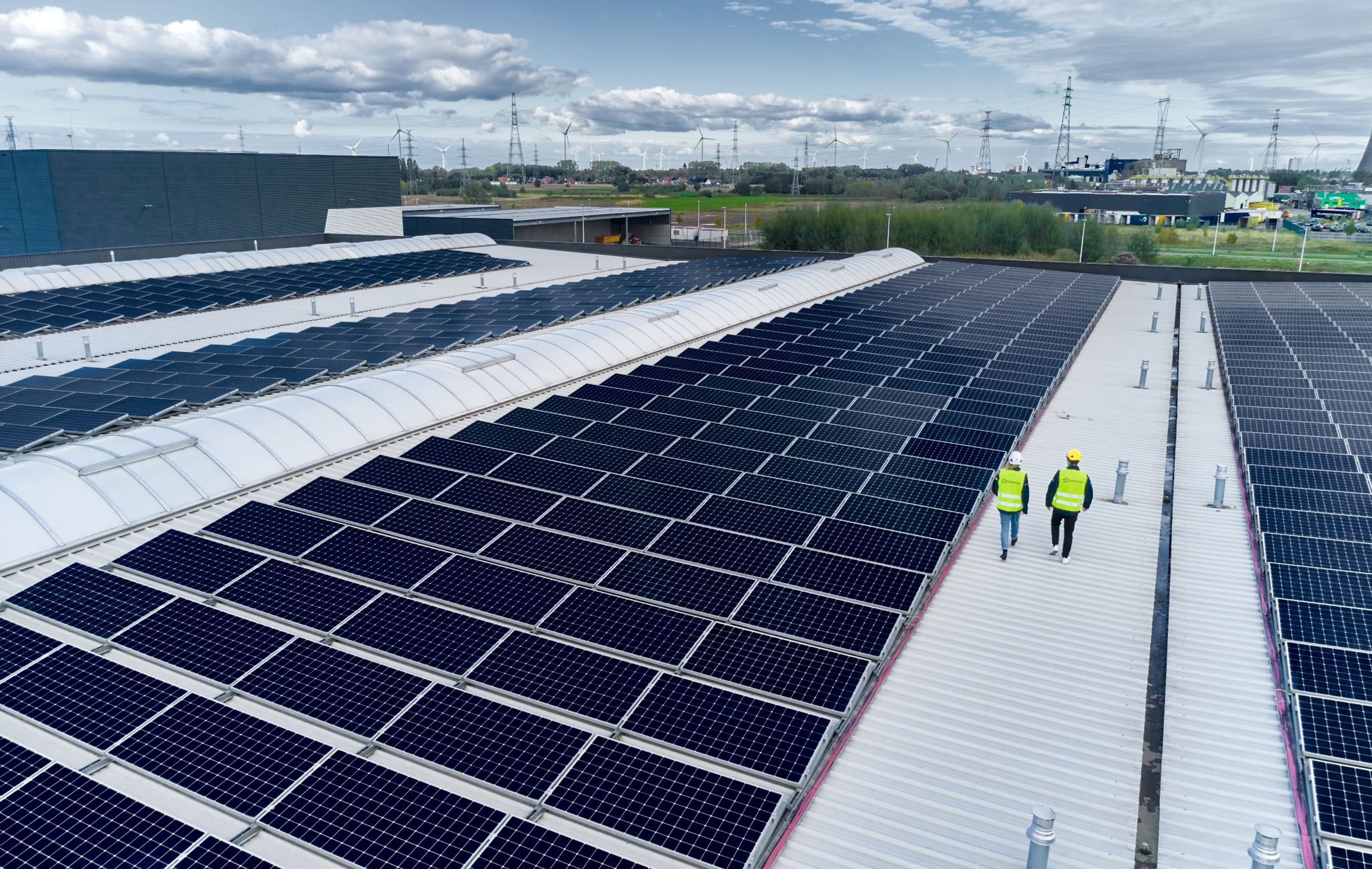 Vue aérienne d’un toit industriel couvert de panneaux solaires, avec deux collaborateurs de Luminus en gilet de sécurité marchant entre les rangées de panneaux
