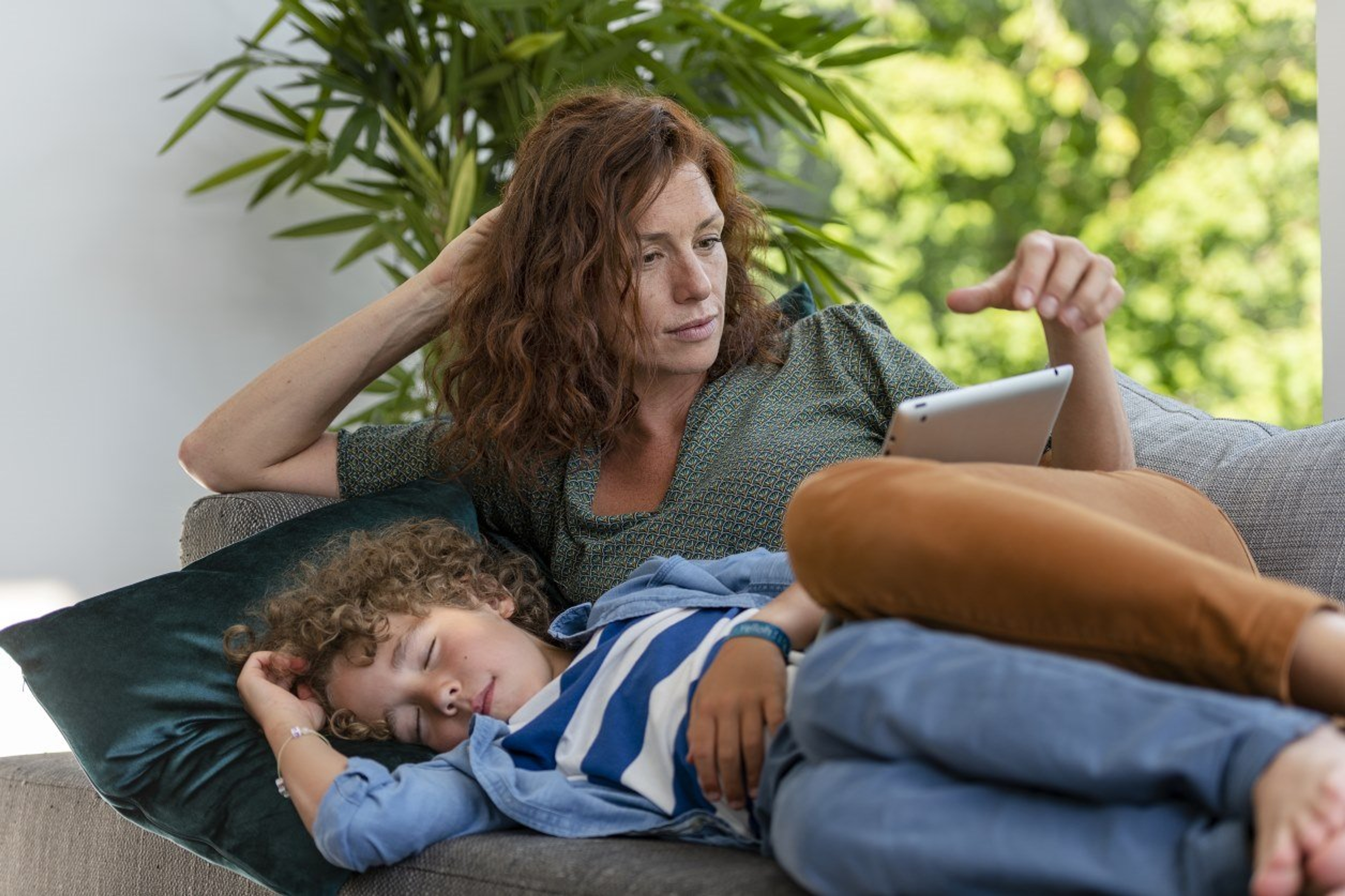 Mama groene top ligt in de zetel en kijkt op tablet. Zoontje met streepjesshirt ligt ernaast te slapen.