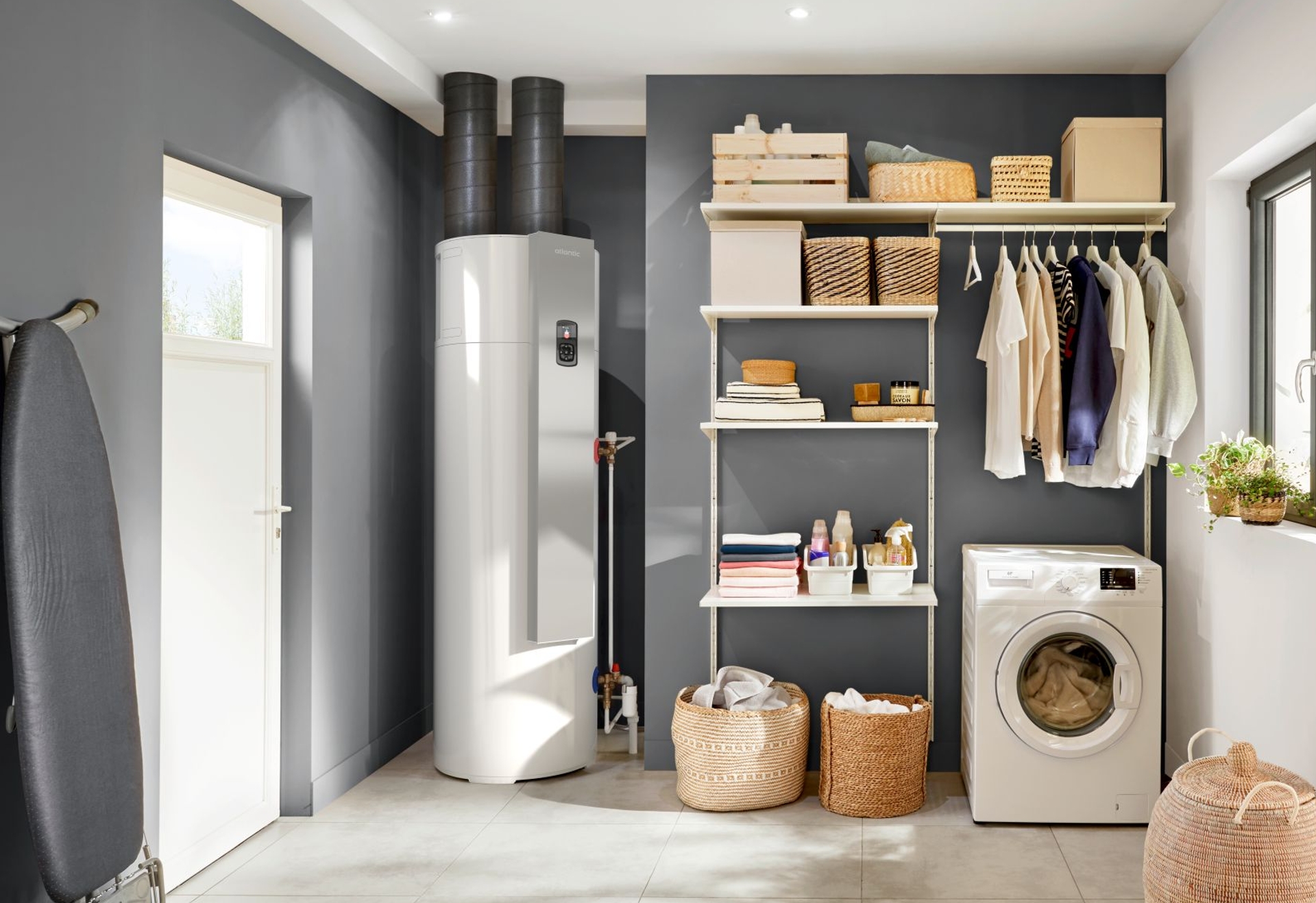 Witte warmtepompboiler in een ruimte met ruimte met wasmachine, wasmanden en rekjes.