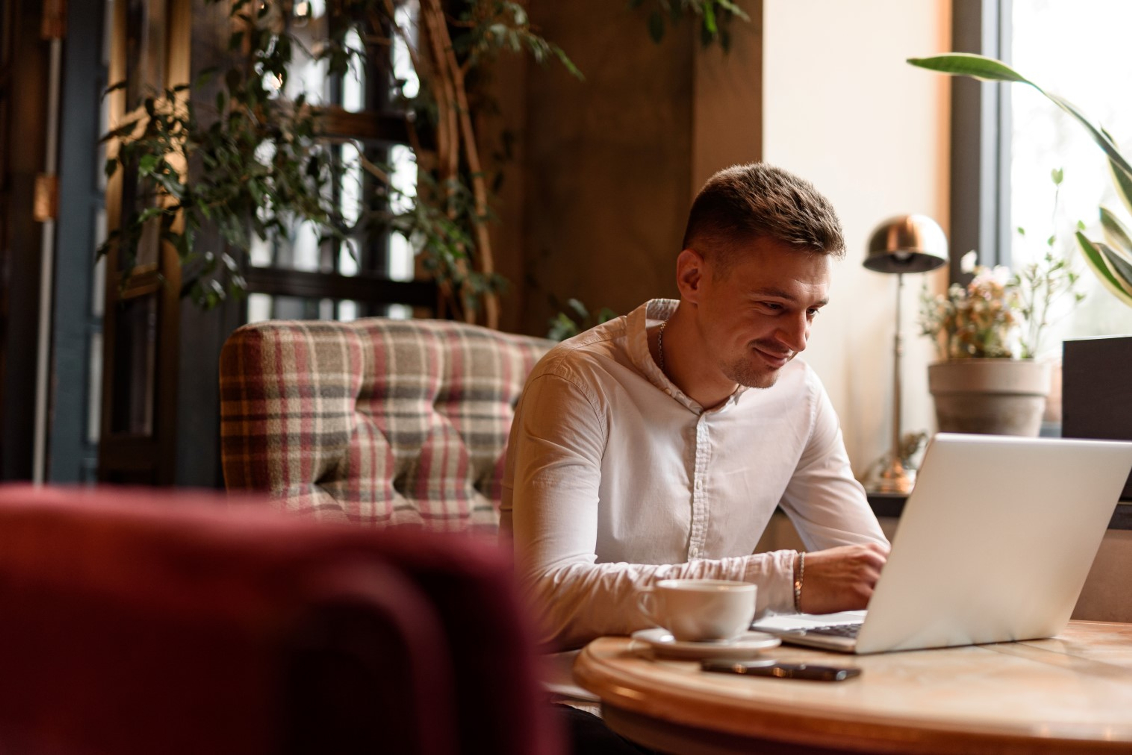 Jonge man kijkt glimlachend op smartphone, geruit kussen achter hem.