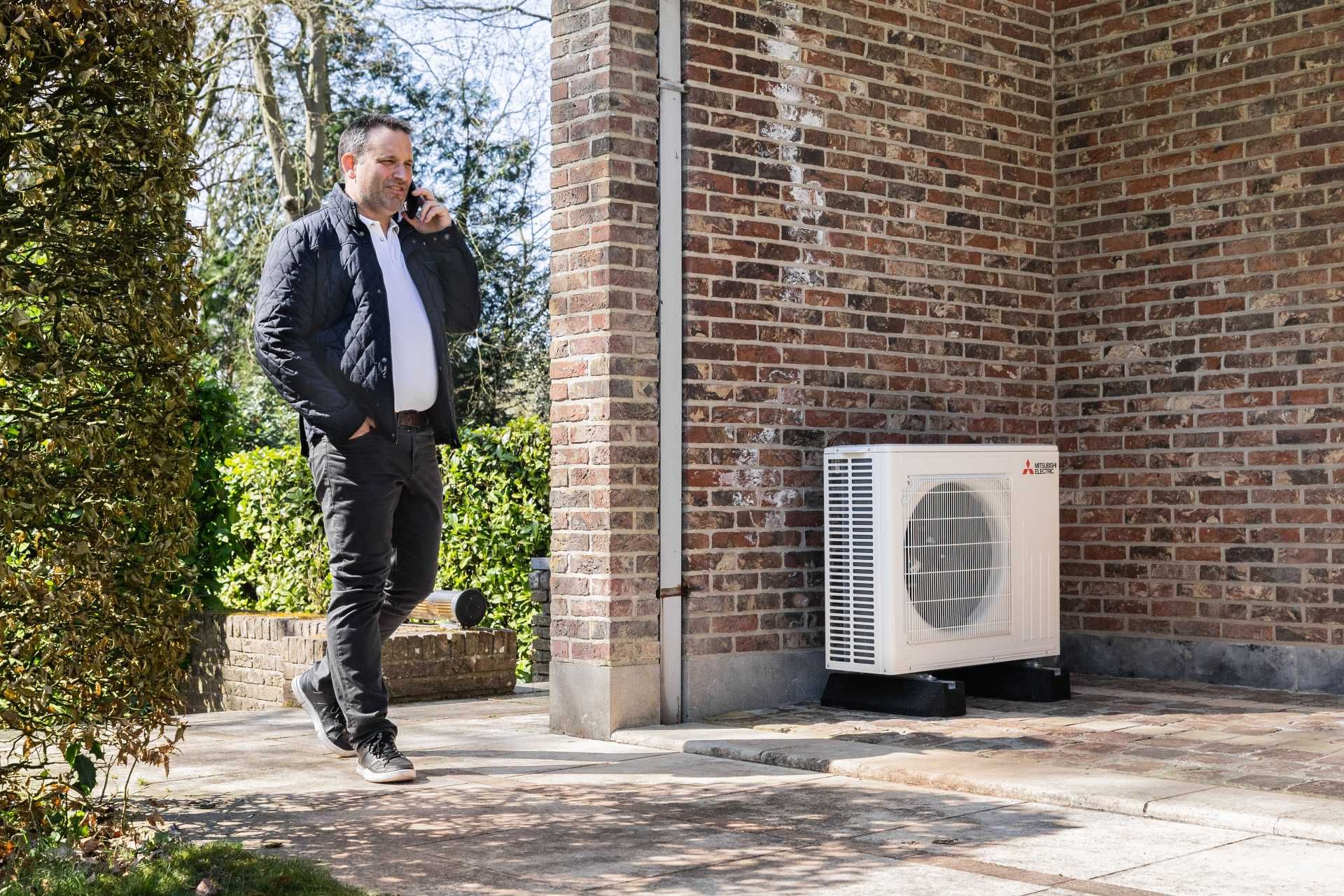 Buitenmuur in baksteen, man met jas aan telefoneert. Er staat een buitenunit tegen de muur.