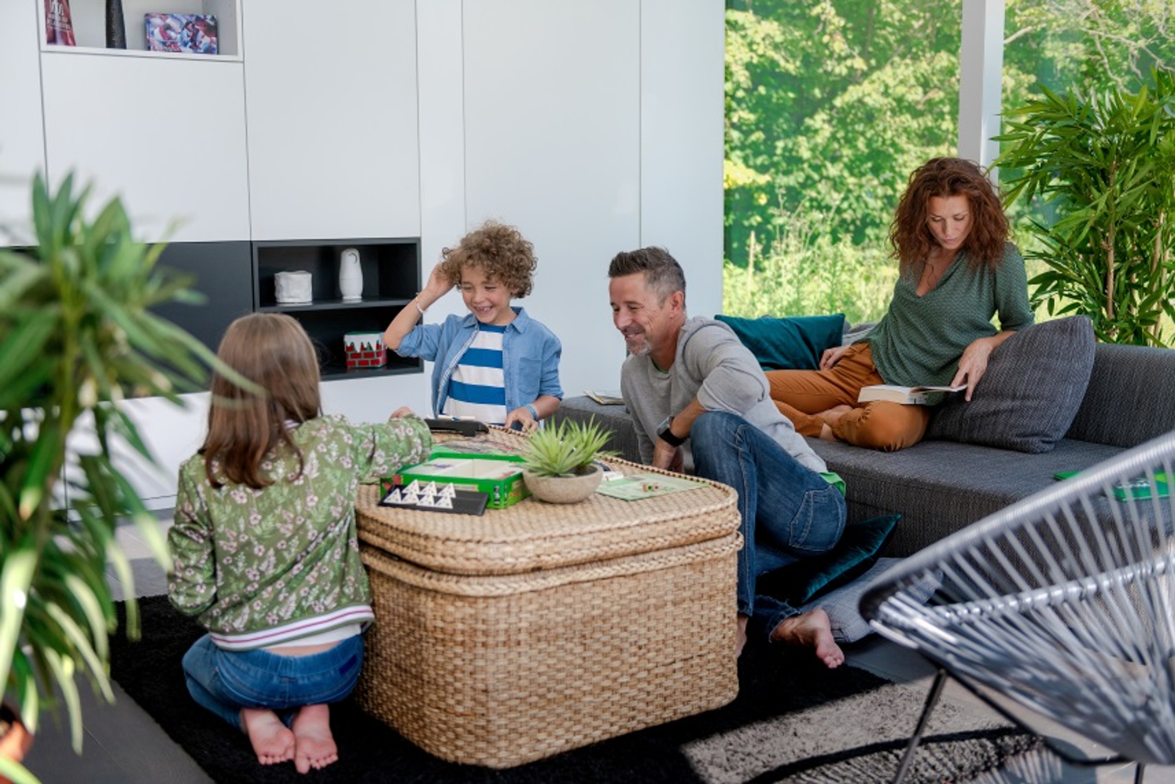 Homme jouant avec 2 enfants sur la table de salon, tandis que la femme lit un livre dan