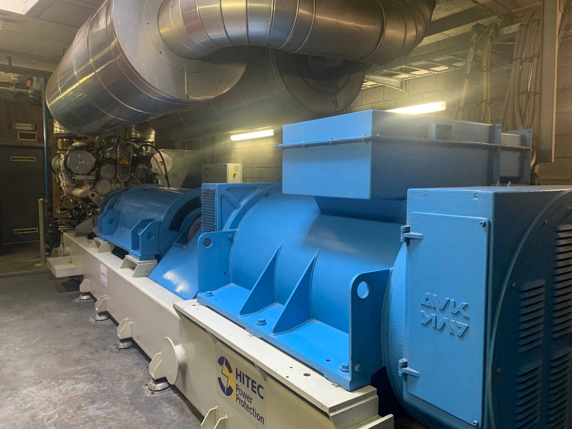 Un grand générateur industriel bleu dans une salle technique, avec des tuyaux apparents et des panneaux de commande visibles