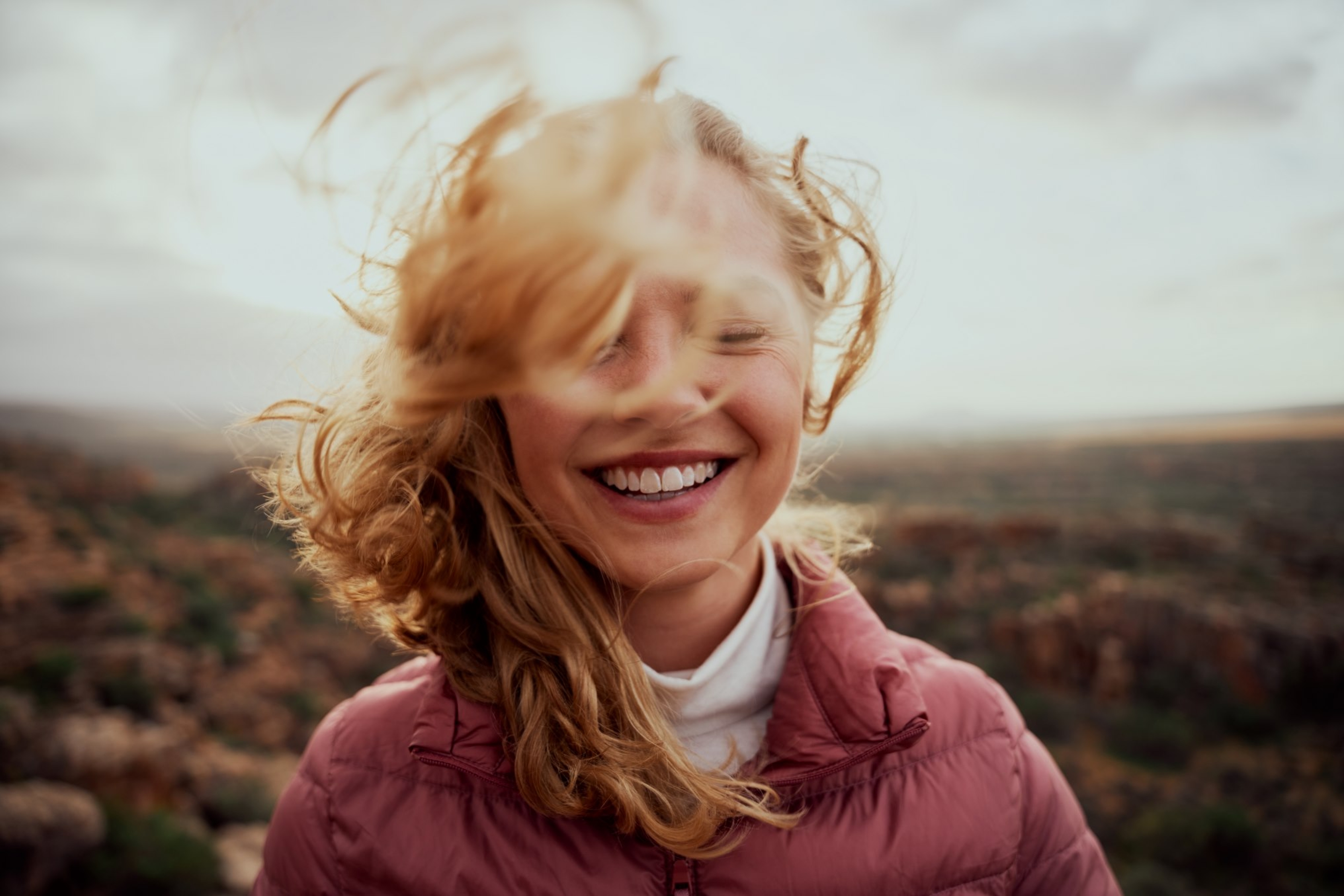 jonge vrouw, lachend, roze jas, blonde haren in de wind in een natuurlandschap