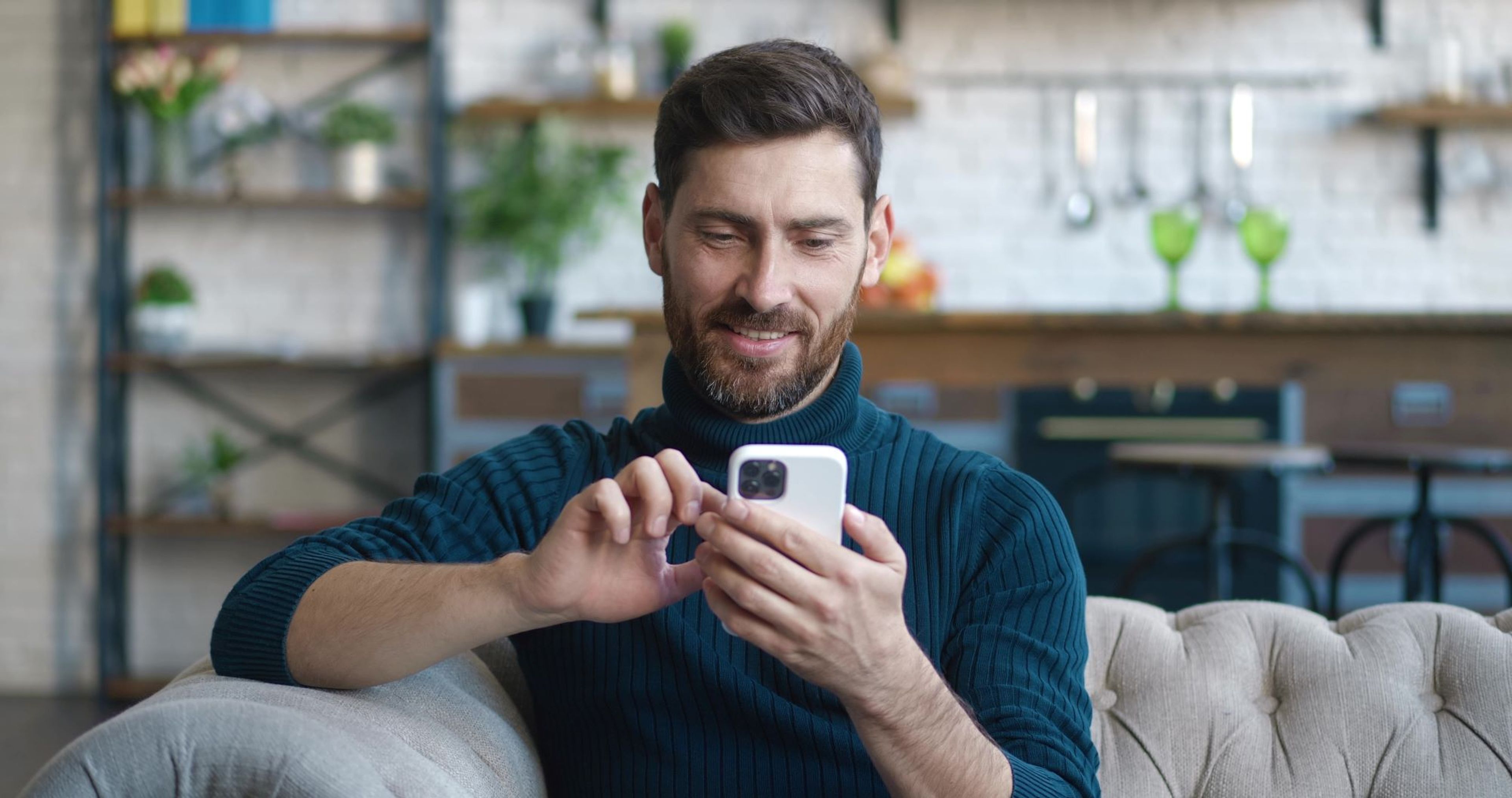 Jonge man met trui en baard zit in de zetel en tikt op zijn smartphone.