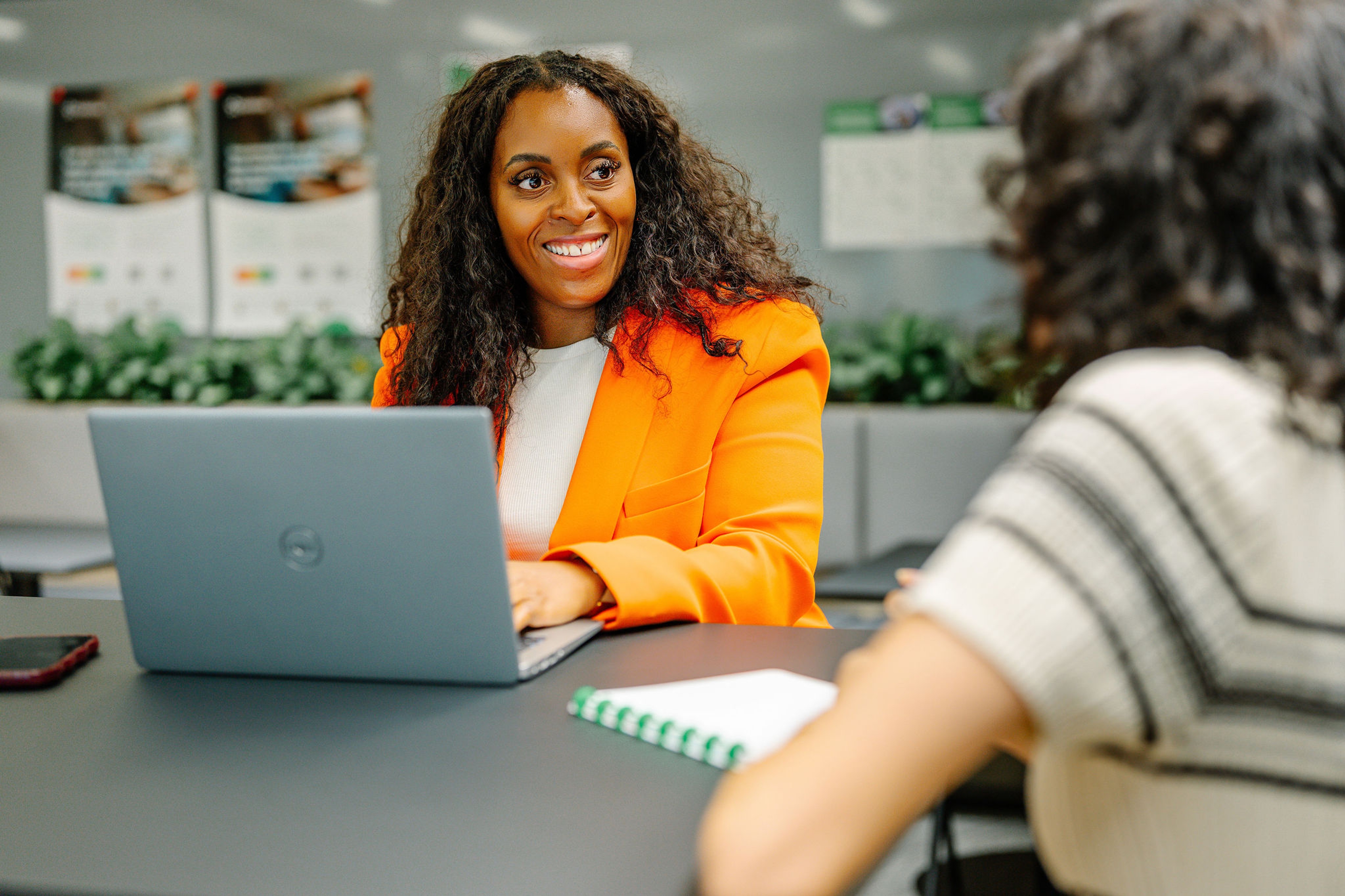 Deux personnes sourient et discutent à une table avec un ordinateur portable et une tasse de café. Deux autres personnes discutent à l'arrière-plan. Le décor est lumineux et rempli de plantes.