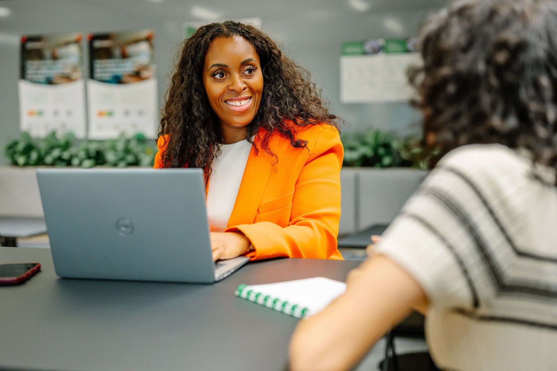 Twee mensen glimlachen en praten aan een tafel met een laptop en een koffiemok. Twee anderen praten op de achtergrond. De omgeving is licht en staat vol met planten.