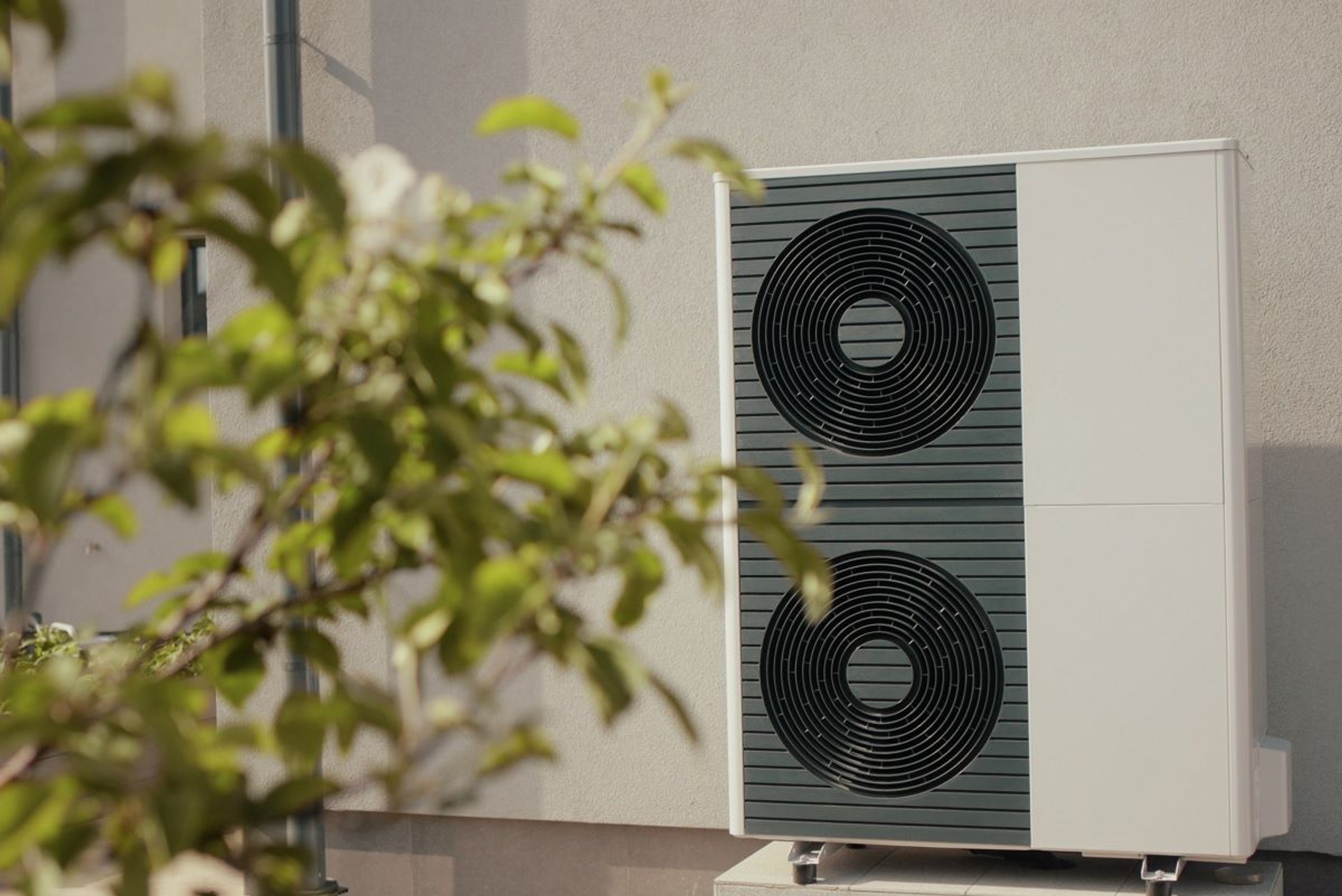Pompe à chaleur hybride avec deux ventilateurs circulaires noirs en façade, installée contre un mur clair, partiellement masquée au premier plan par des branches feuillues