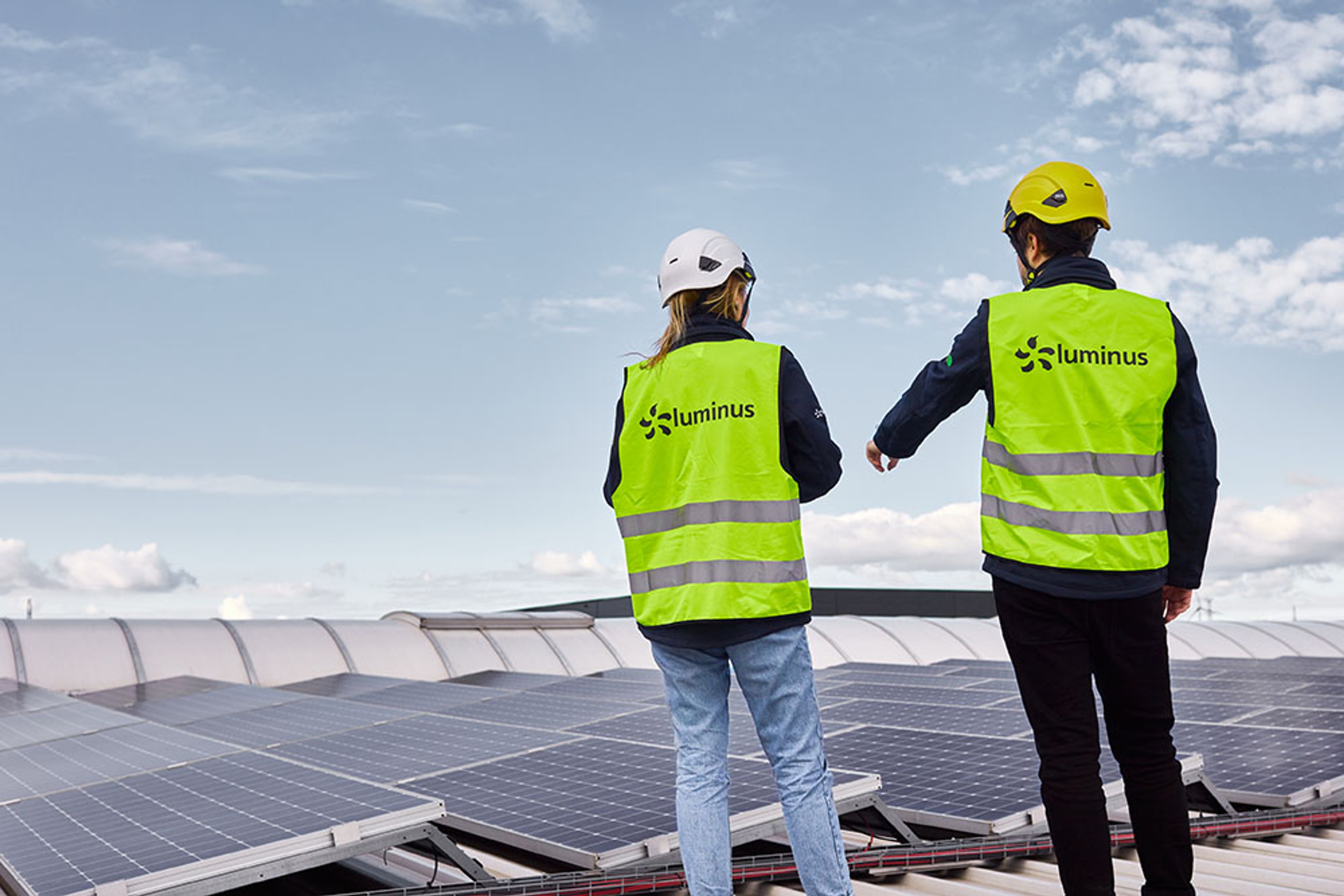 Vrouw en man met helm op en gele hesjes van Luminus aan staan op een dak vol zonnepanelen.