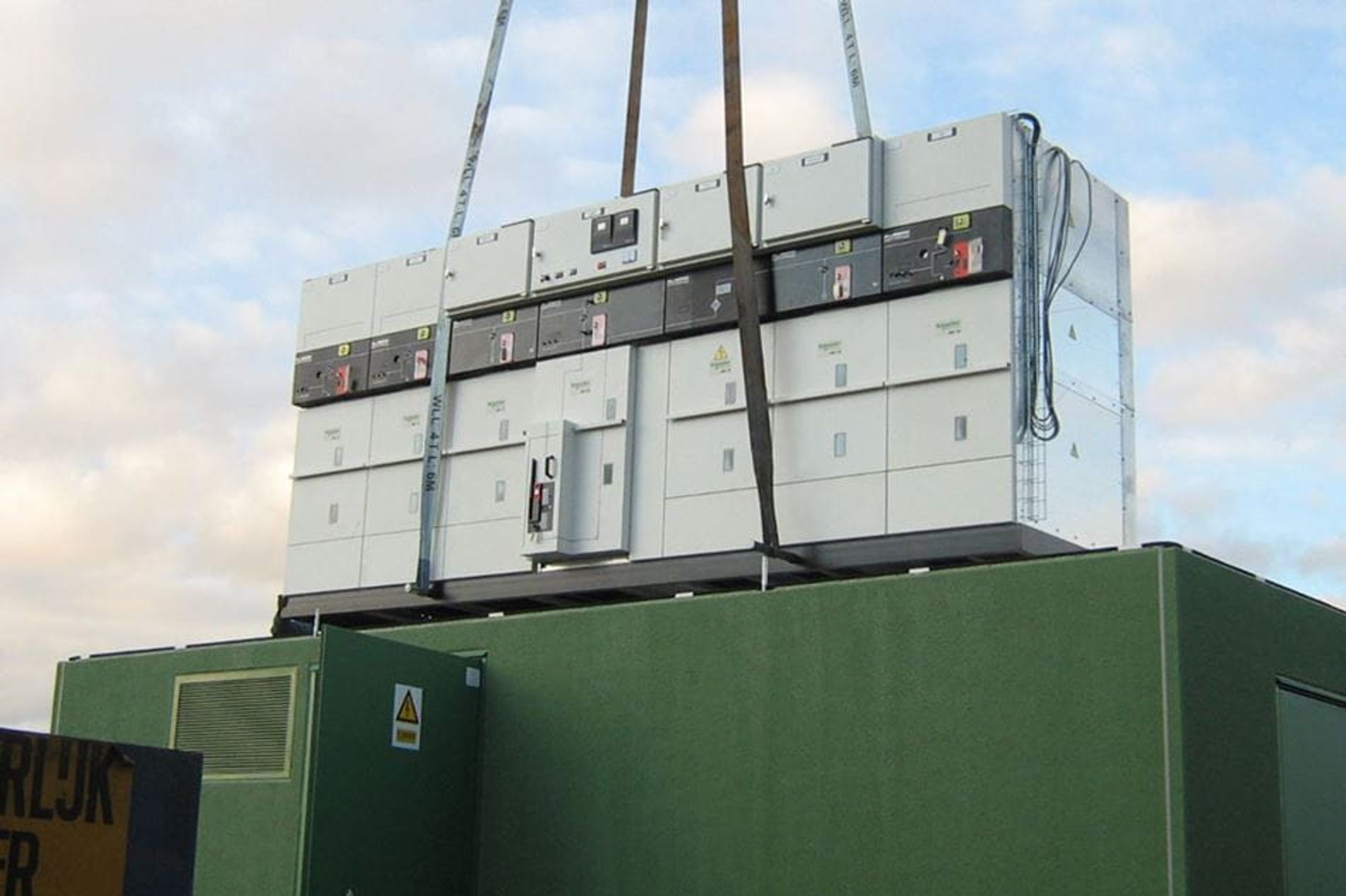 Een grote hoogspanningscabine wordt met hijsbanden door een kraan in een groene container geplaatst.getild. 