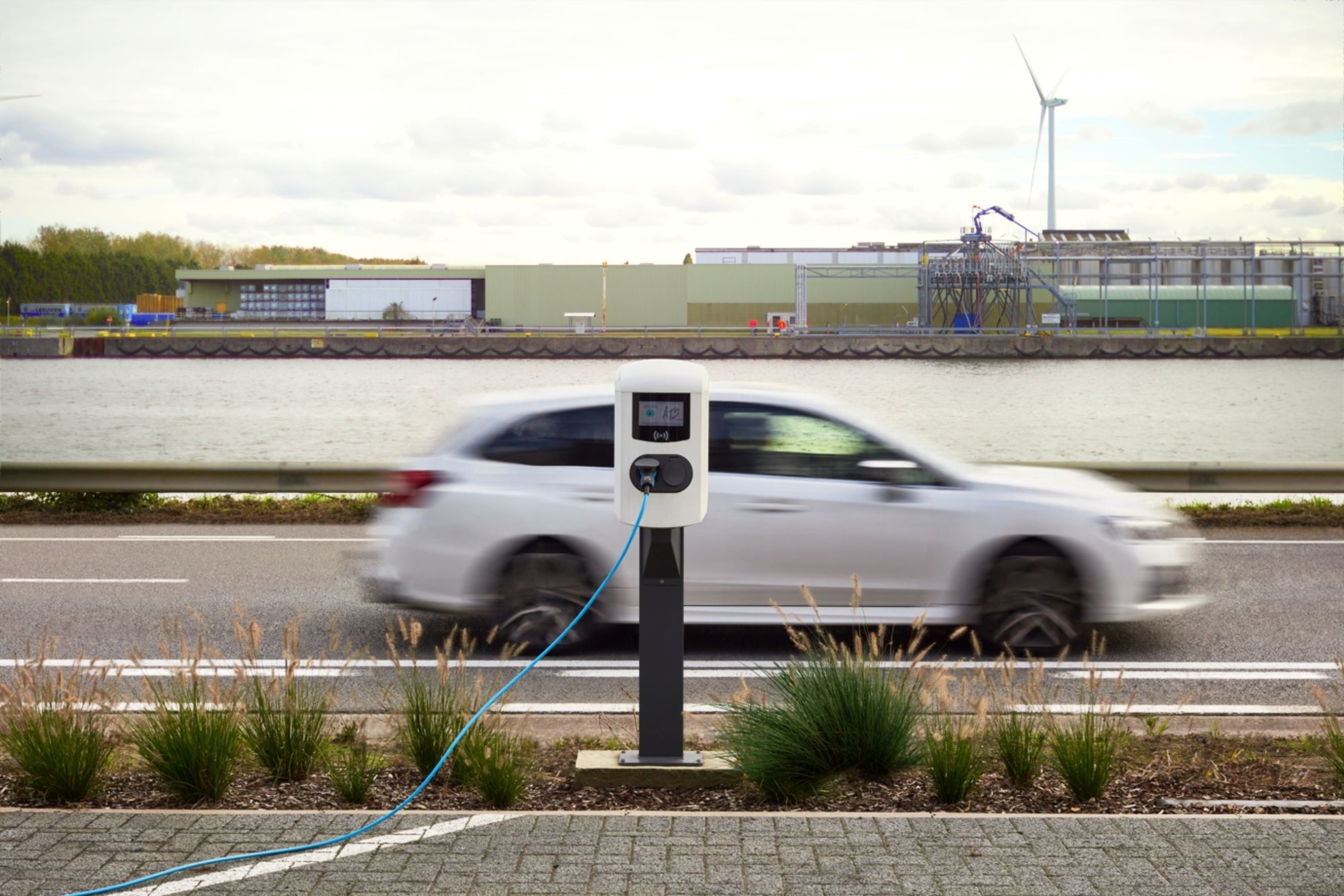 Witte publieke laadpaal en we zien een wazige auto daarachter voorbijrijdt.