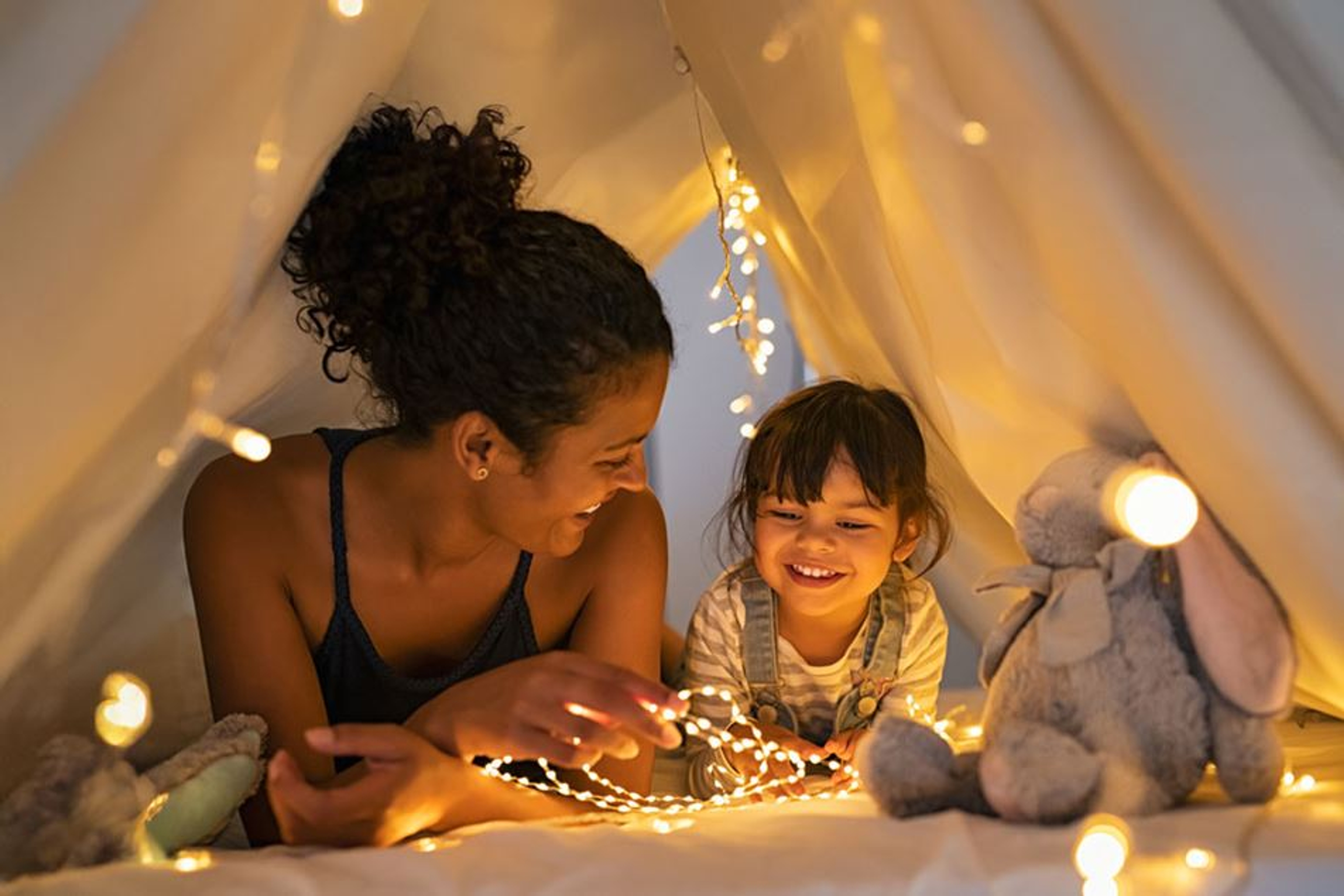 Jeune femme et petite fille couchées sous une tente blanche, entourées de guirlandes lumineuses et de peluches