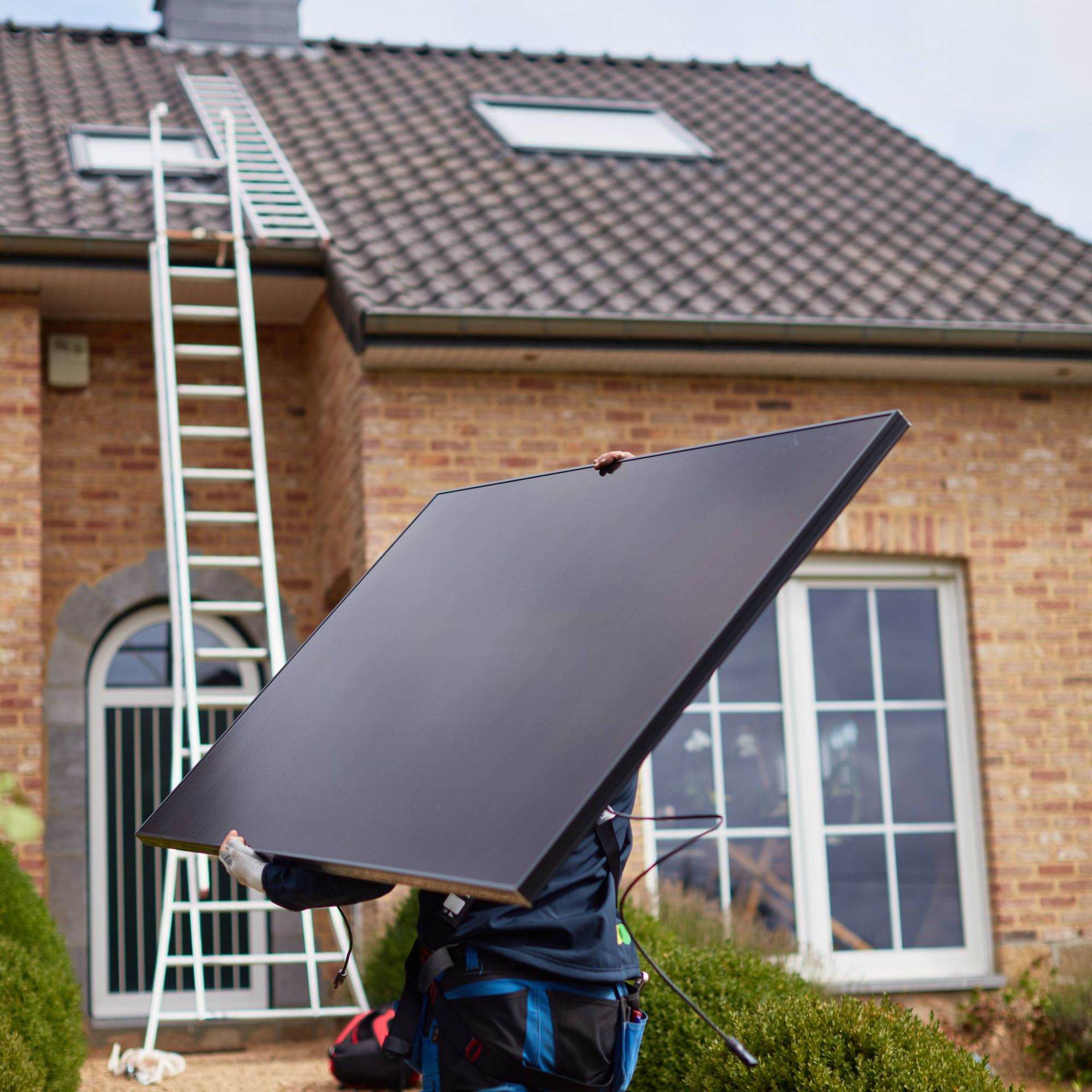 Installateur van zonnepanelen draagt een paneel op de schouder richting ladder en dak van een huis