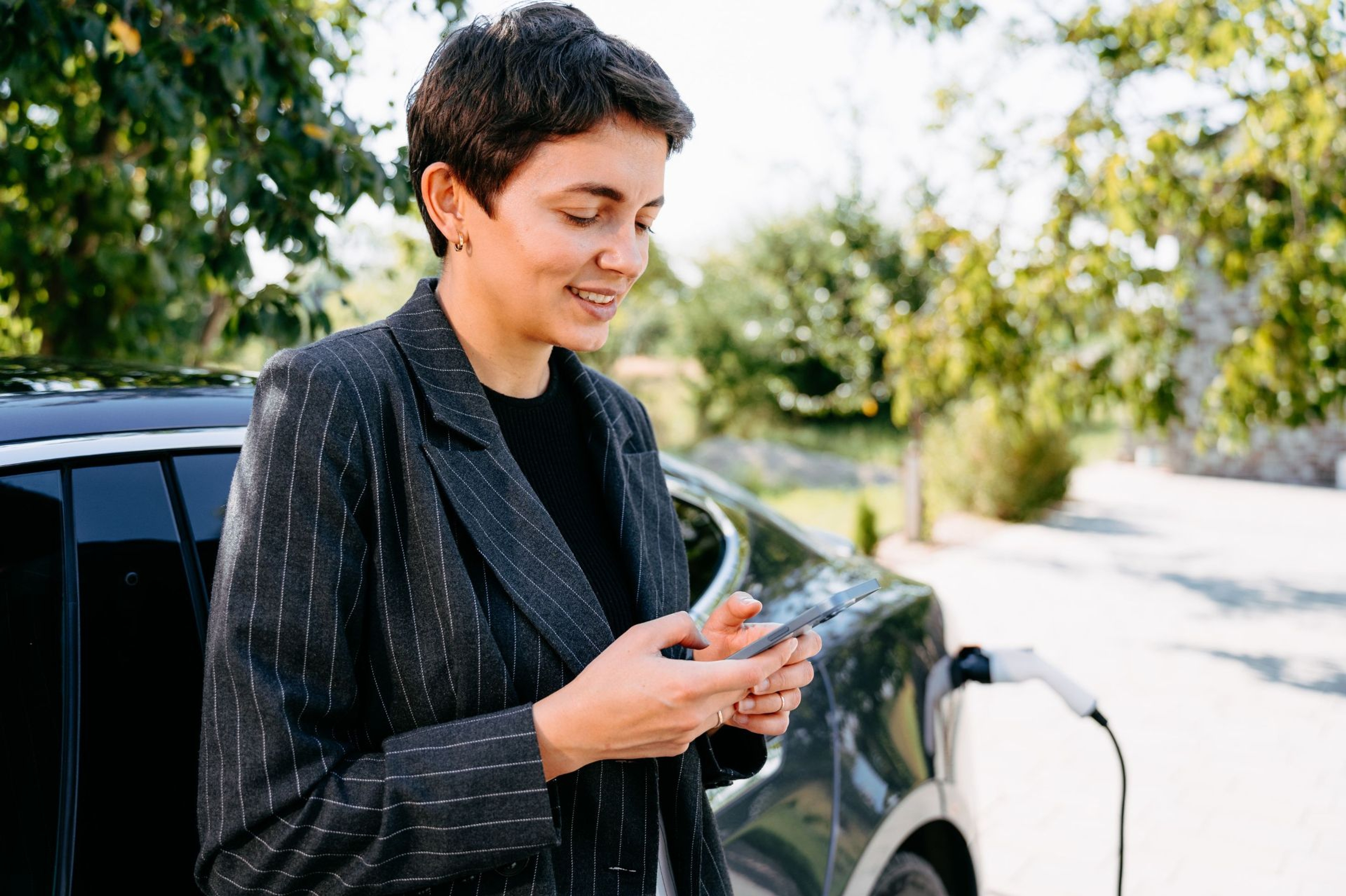 Jonge business vrouw met smartphone in de hand bij haar opladende elektrische wagen