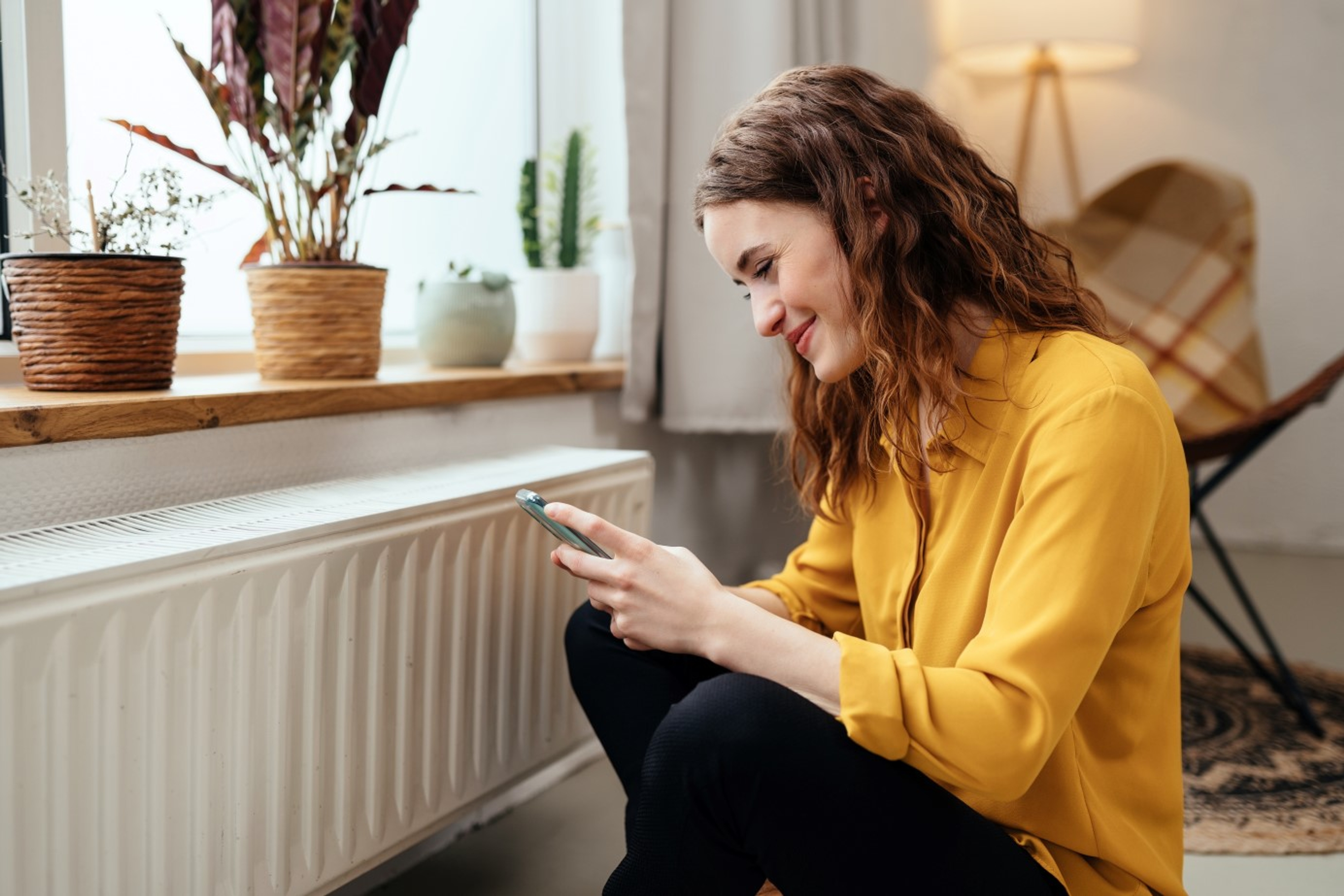 Jonge vrouw met gele trui en smartphone in de hand zit in woonkamer bij de radiator