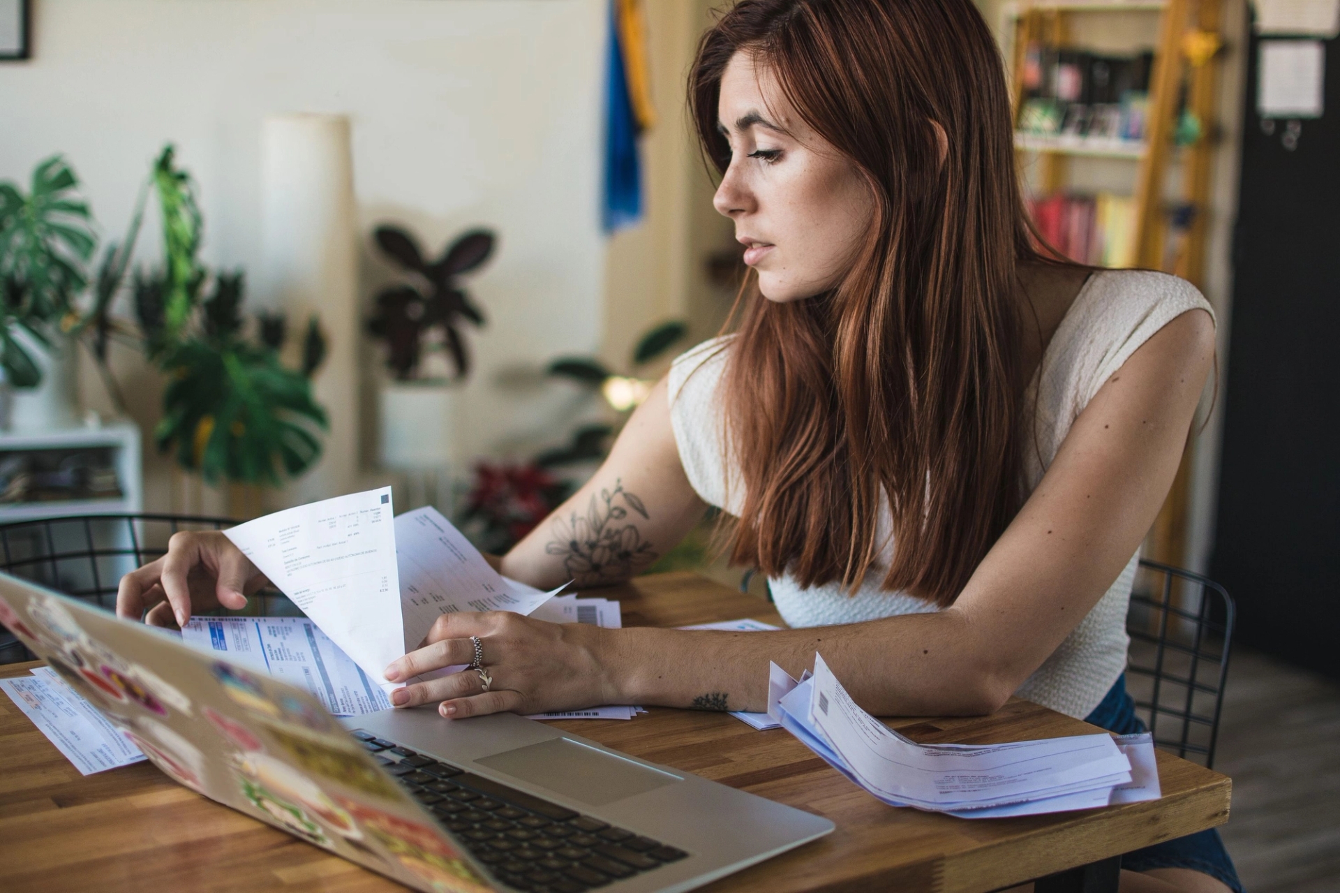 Jeune femme rousse assise à une table, concentrée sur des papiers et factures qu’elle consulte devant un ordinateur portable, dans un intérieur lumineux avec des plantes