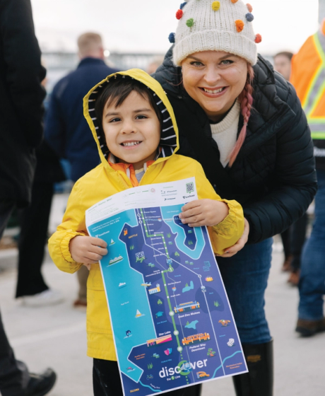 A child in a yellow raincoat and adult smiling while holding a Link Light Rail "discover" route map at the opening event