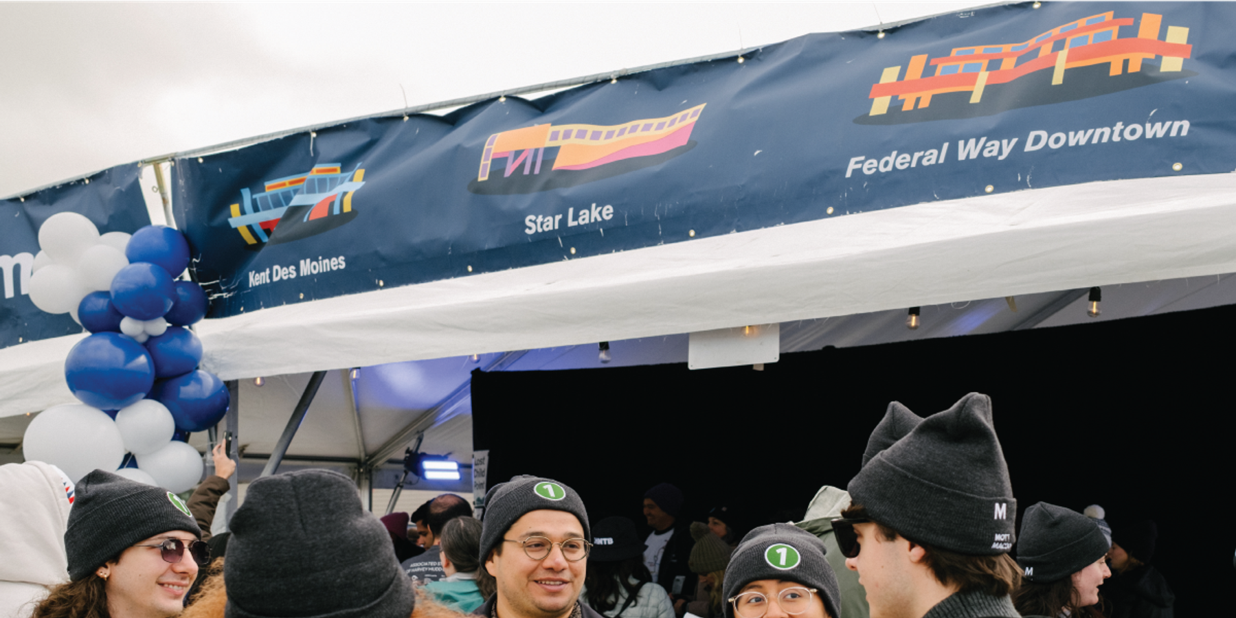 Sound Transit event staff in branded beanies under a blue banner showing Kent Des Moines, Star Lake, and Federal Way Downtown stations