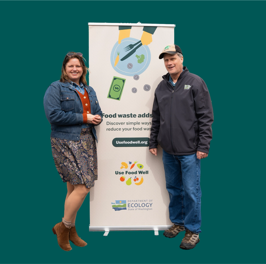 Two campaign representatives pose beside a Use Food Well pull-up banner reading "Food waste adds up" on a teal background