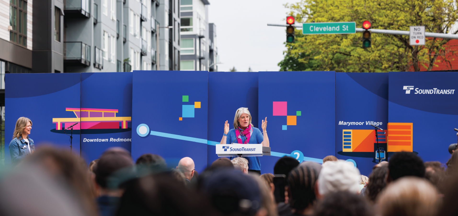 Speaker addresses a crowd at the Sound Transit Link Light Rail opening ceremony in front of a blue branded backdrop showing station stops