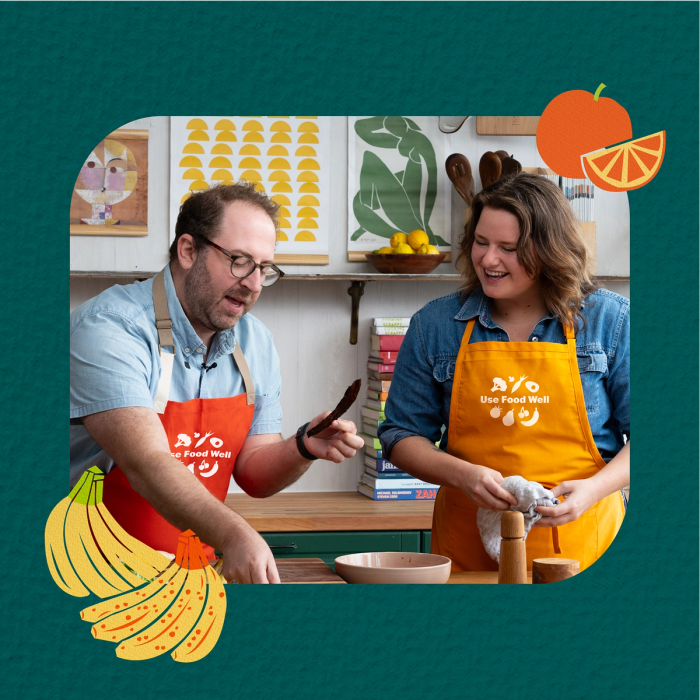 Two people wearing Use Food Well aprons cooking together, with illustrated fruit decorations on a teal background