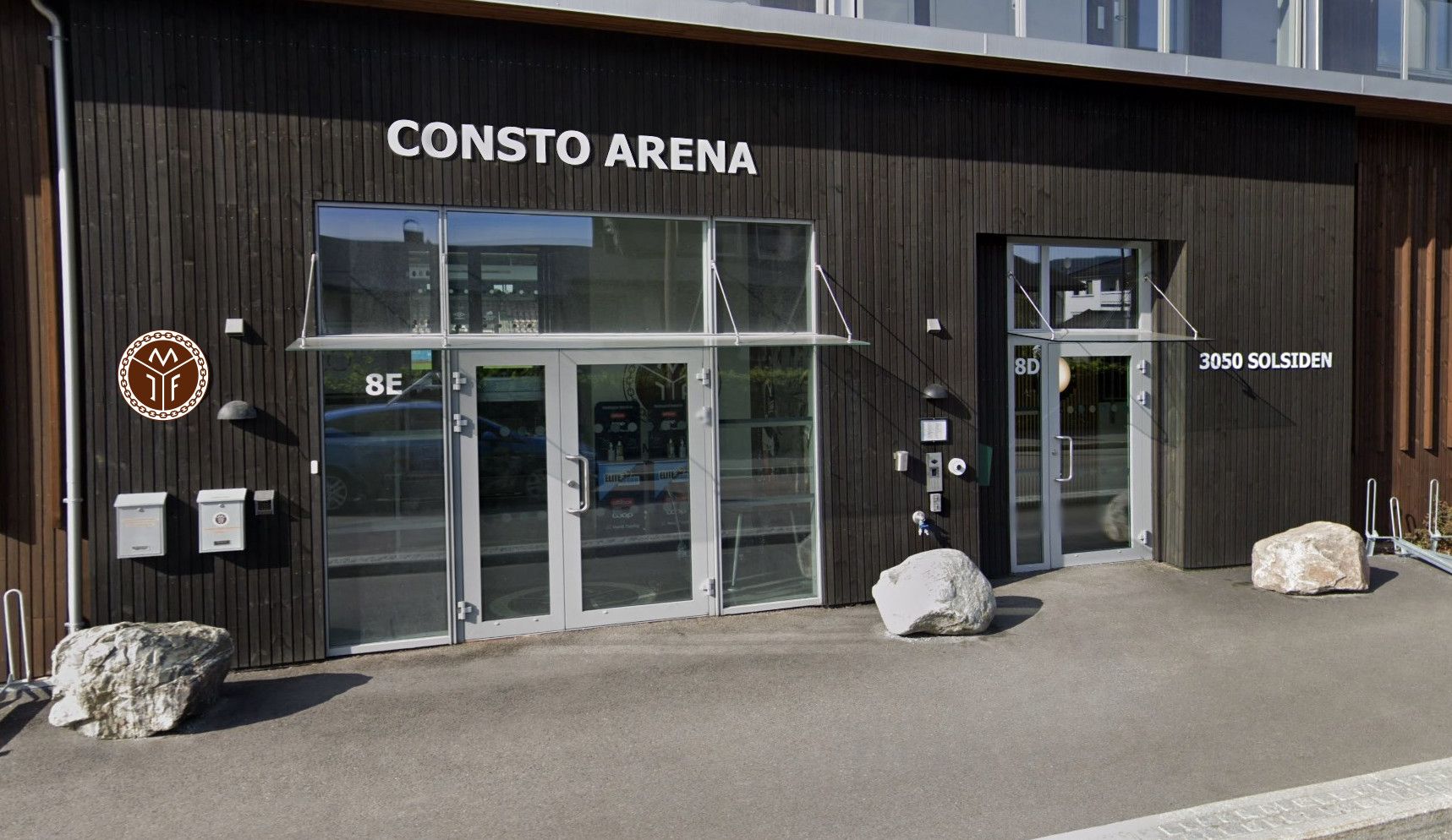 Inngangen til Consto Arena i Mjøndalen, en mørk trefasade med Mjøndalen IFs logo ved døra.