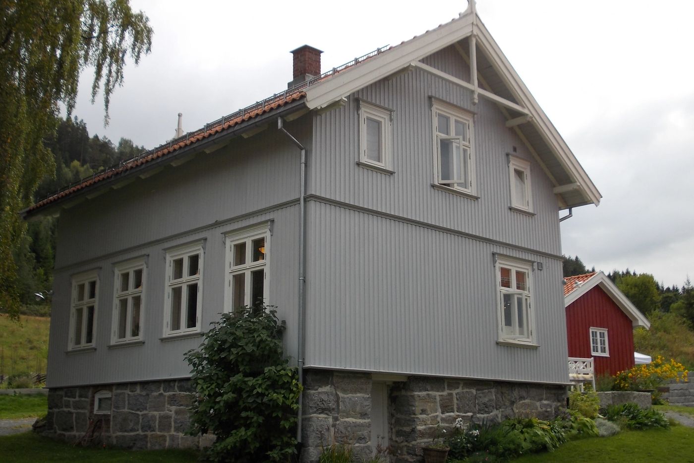Hovedhuset på Portåsen, et grått trehus i to etasjer med hage foran og et rødt uthus i bakgrunnen.