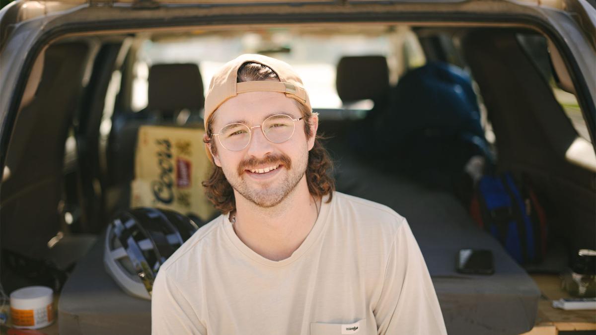 A moustachioed Mo Bro, with spectacles and a backwards baseball cap, poses for a photo sitting in the open trunk of his car