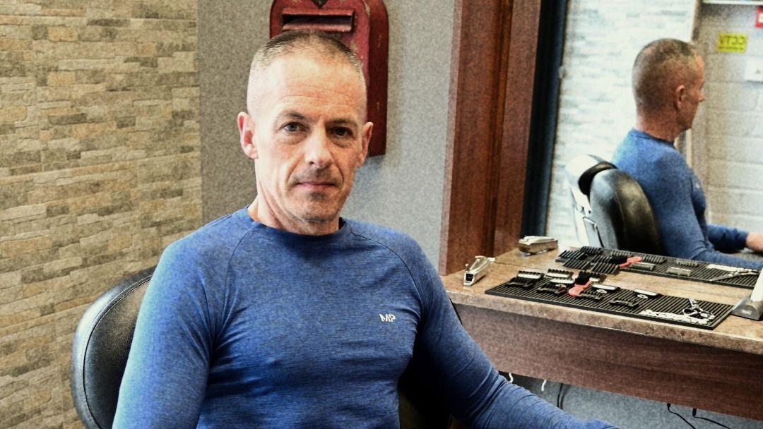 David sat in a chair in a barbers looking at camera