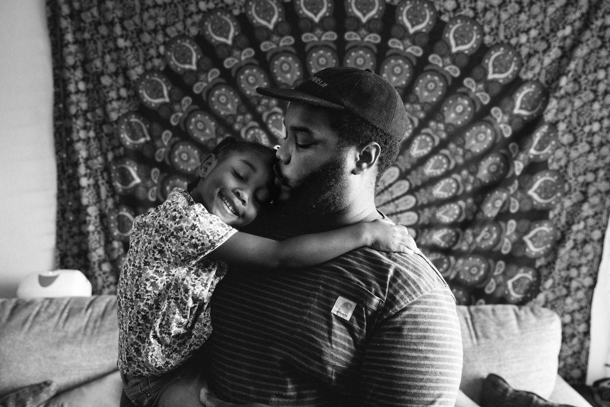 A father kisses his young daughter on the cheek as she smiles and hugs him.