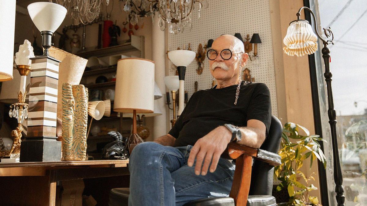 A man sits in a barber chair in an antique shop, he's sporting large-framed black glasses, and a perfectly groomed, bushy, white, moustache