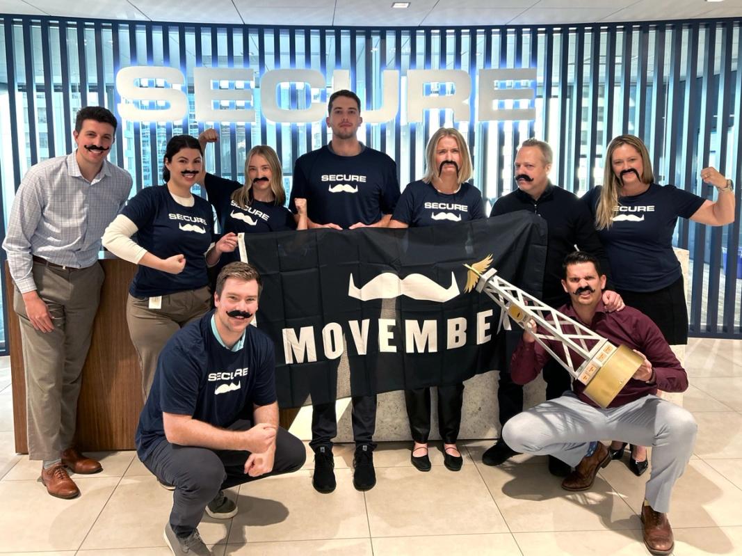 A group of Movember fundraisers are posing for the camera and smiling, holding and wearing Movember merch