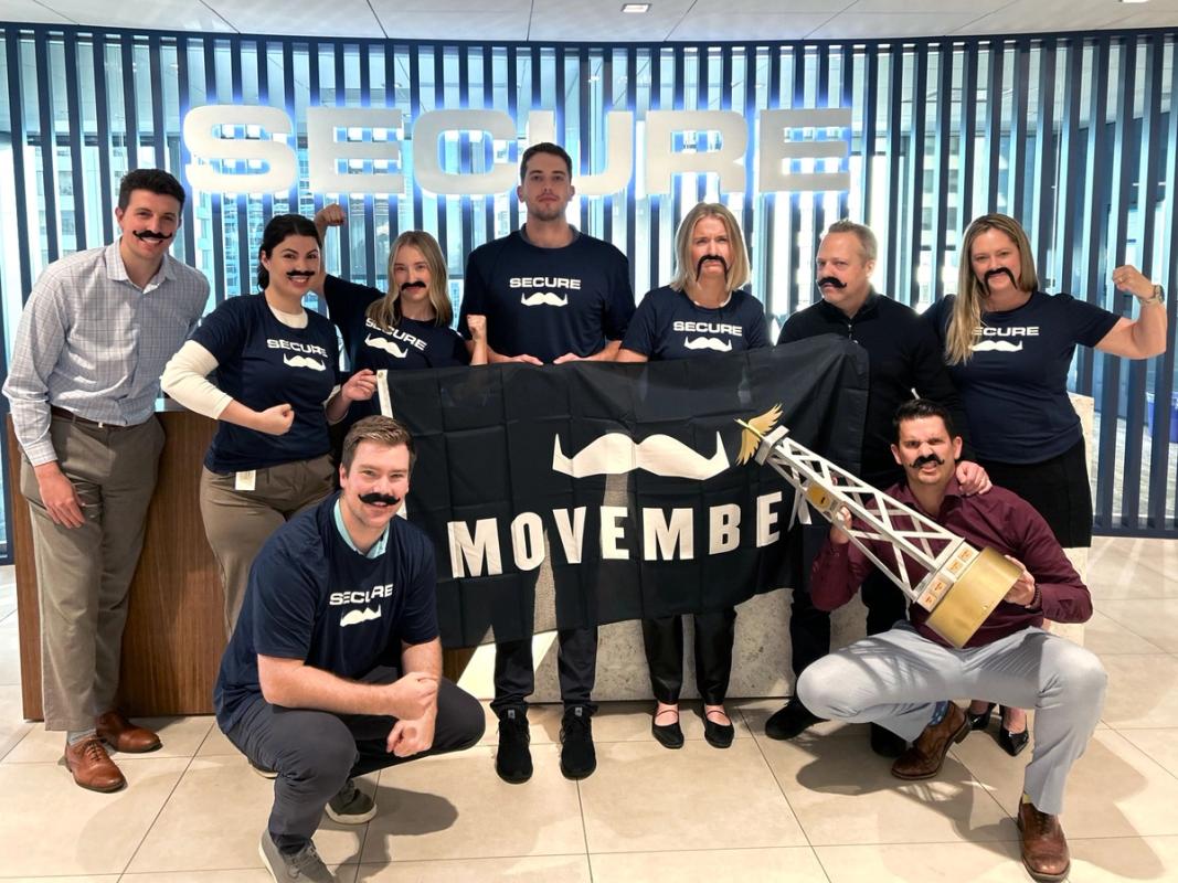 A group of Movember fundraisers are posing for the camera and smiling, holding and wearing Movember merch