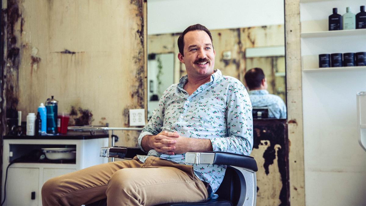 A man in a blue, patterned, button up shirt poses in a barber chair in an LA barbershop