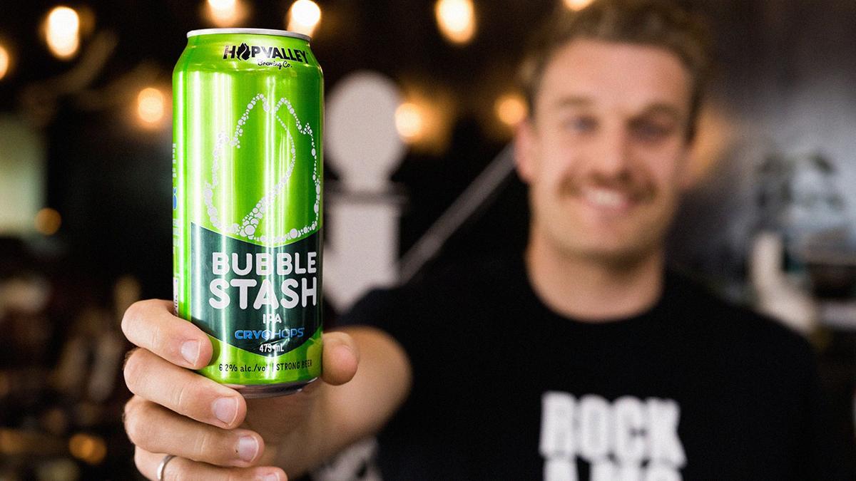 A tall can of beer that reads "Hop Valley Bubble Stash" is held in a forefront by a man