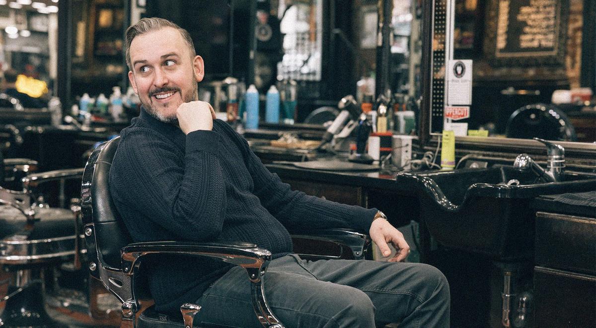 Man sitting in a barber chair grinning with his hand on his chin and looking over his shoulder.