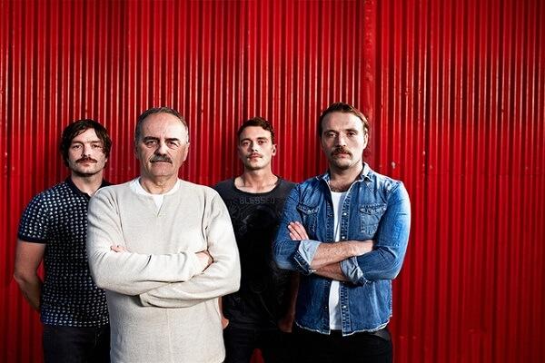 Four men of different ages standing together in front of a red wall.