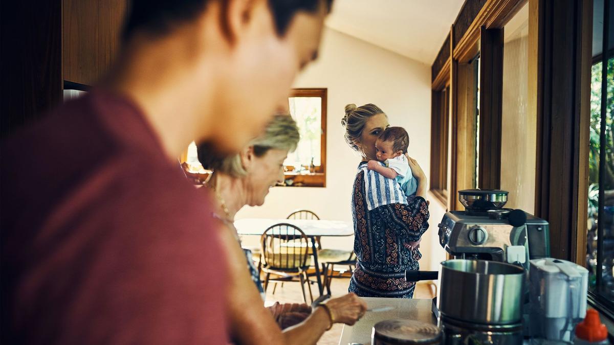 Mother holds baby in kitchen as father and grandmother make food.