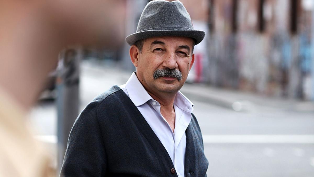 A candid photo of a moustachioed man standing on the pavement 