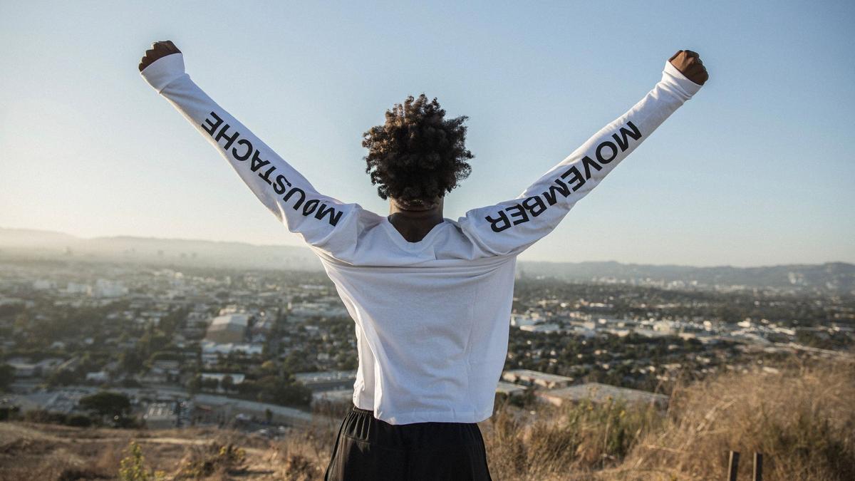 A young man stands with his arms raised over a valley. The sleeves of his shirt say "Movember" "Moustaches"