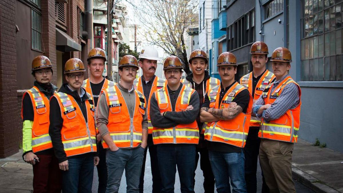 Group of men with high vis jackets and hard hats pose down alley way looking at camera 