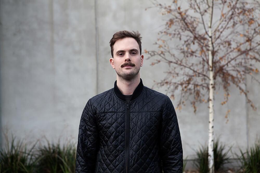 A young man with a moustache stands outdoors in a black jacket, looking directly at the camera with a neutral expression.